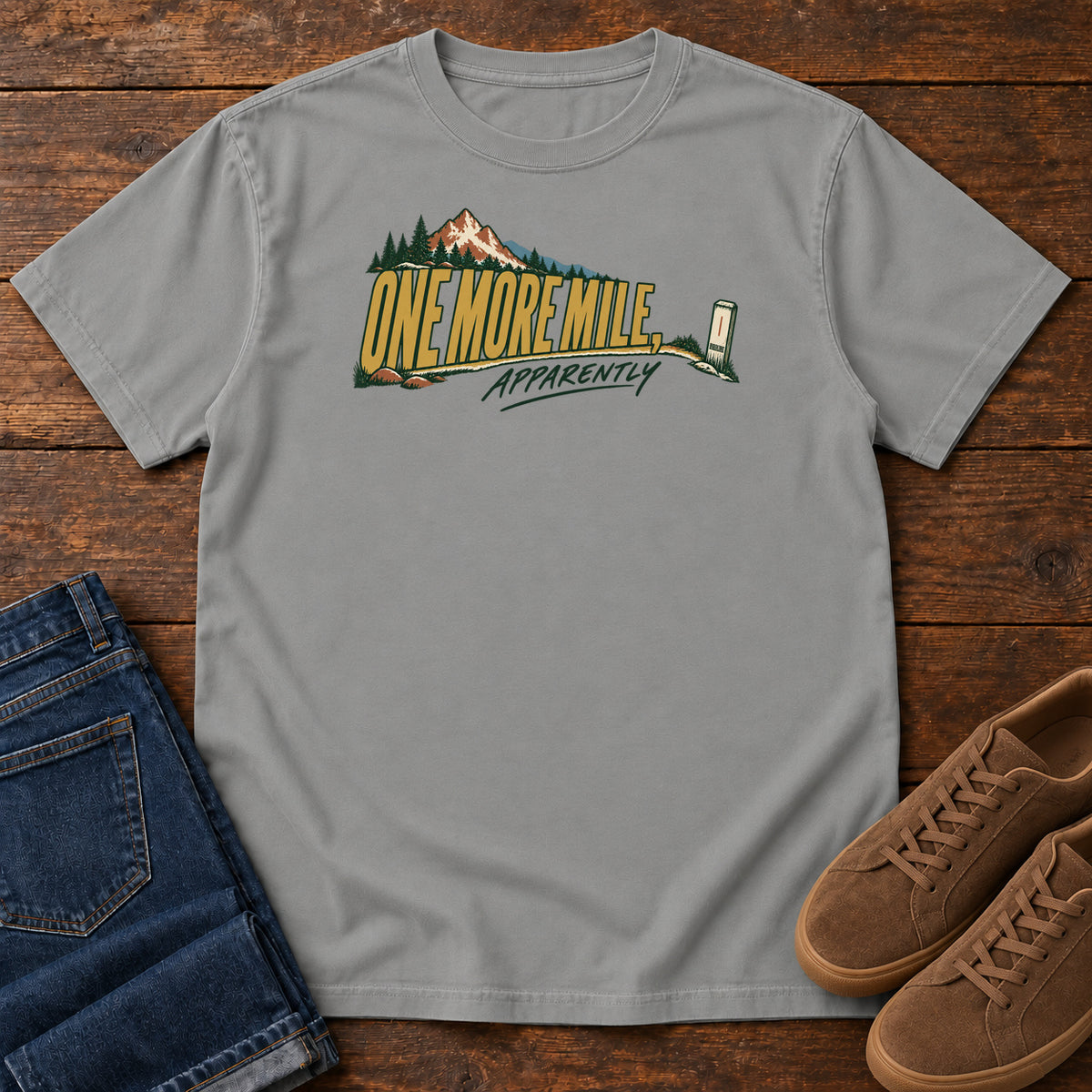 One More Mile Apparently T-Shirt in Grey, front laid flat on wood, featuring gold slogan lettering with a mountain, pine trees and a small trail marker.