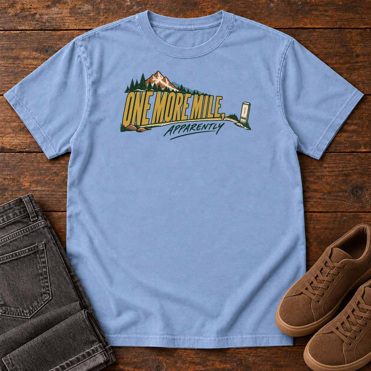 One More Mile Apparently T-Shirt in Washed Denim, front laid flat on wood, featuring gold slogan lettering with a mountain, pine trees and a small trail marker.
