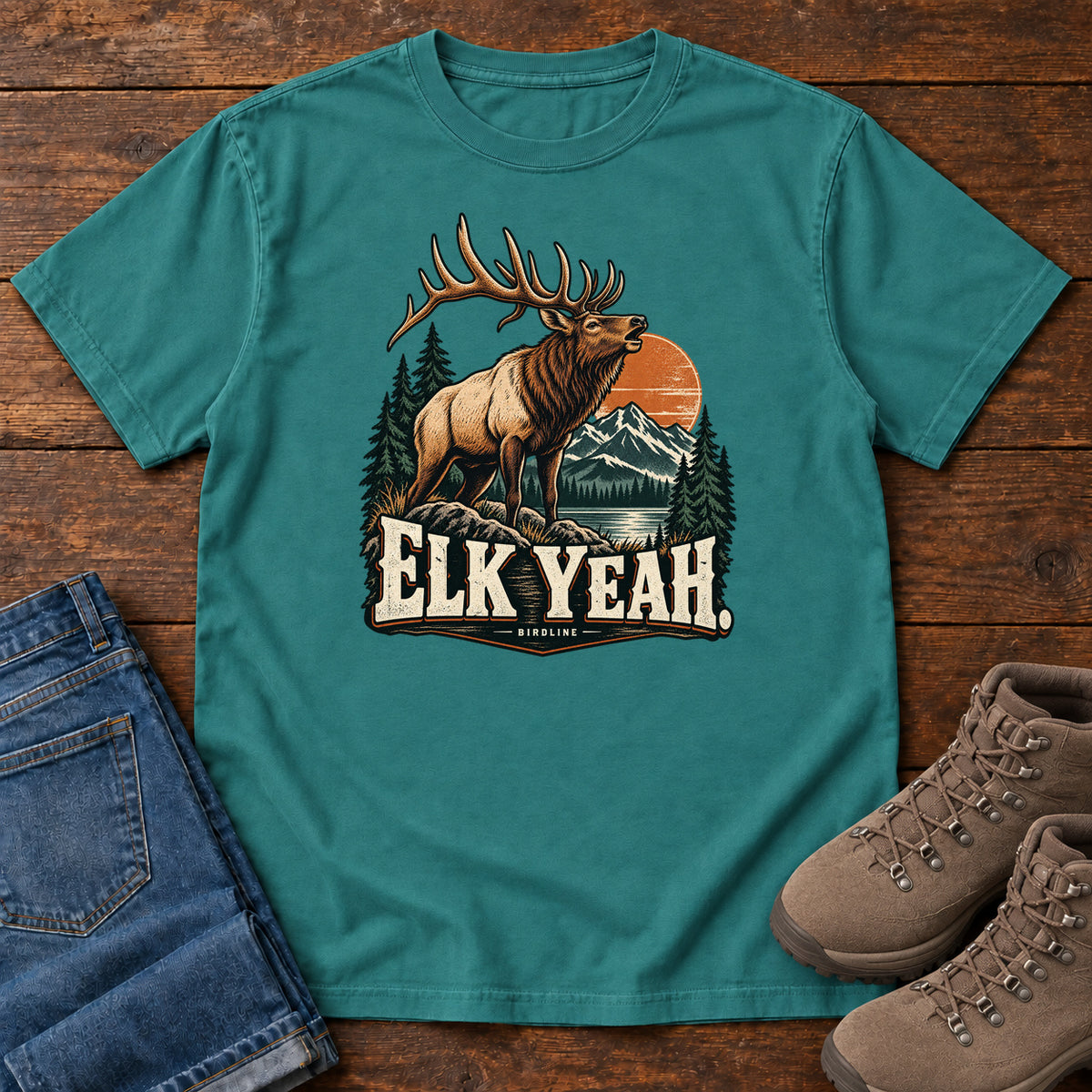 Elk Yeah T-Shirt in Emerald, front laid flat on wood, featuring an antlered elk before mountains, pine trees and an orange sun, above cream lettering.