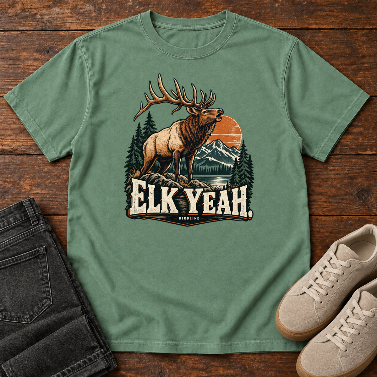 Elk Yeah T-Shirt in Light Green, front laid flat on wood, featuring an antlered elk before mountains, pine trees and an orange sun, above cream lettering.