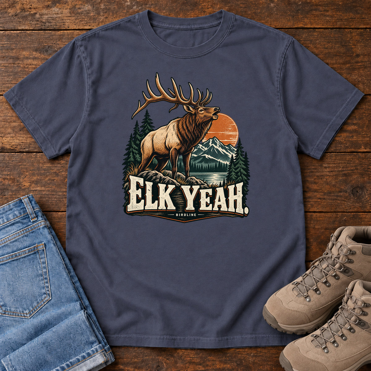 Elk Yeah T-Shirt in Navy, front laid flat on wood, featuring an antlered elk before mountains, pine trees and an orange sun, above cream lettering.