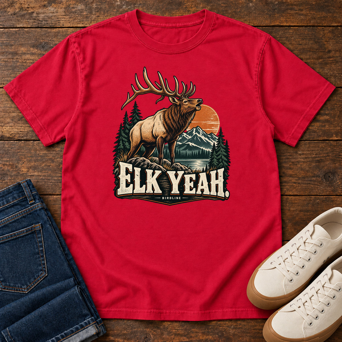 Elk Yeah T-Shirt in Red, front laid flat on wood, featuring an antlered elk before mountains, pine trees and an orange sun, above cream lettering.