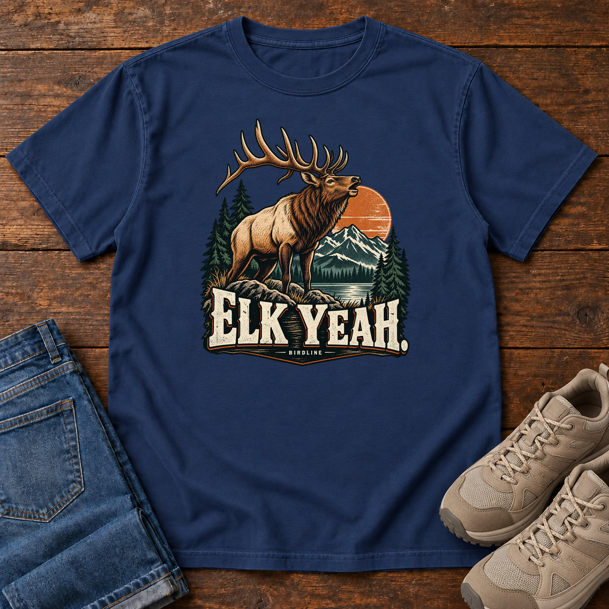 Elk Yeah T-Shirt in True Navy, front laid flat on wood, featuring an antlered elk before mountains, pine trees and an orange sun, above cream lettering.