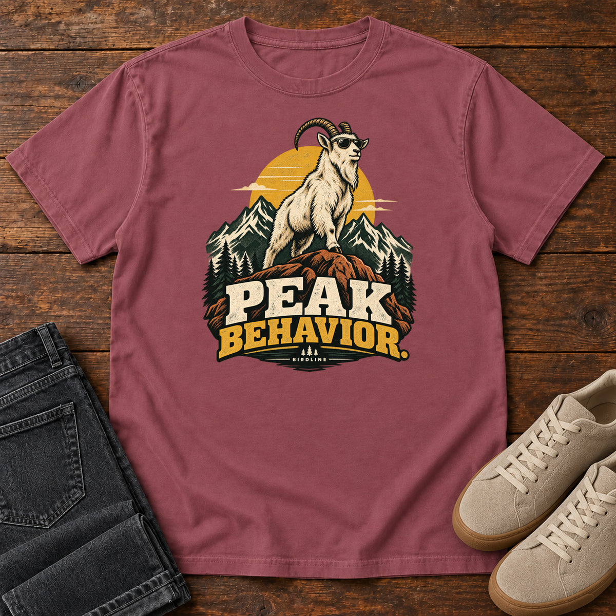 Peak Behavior T-Shirt in Brick, front laid flat on wood, featuring a horned white goat wearing sunglasses on a rocky peak, with mountains, a gold sun and bold lettering.