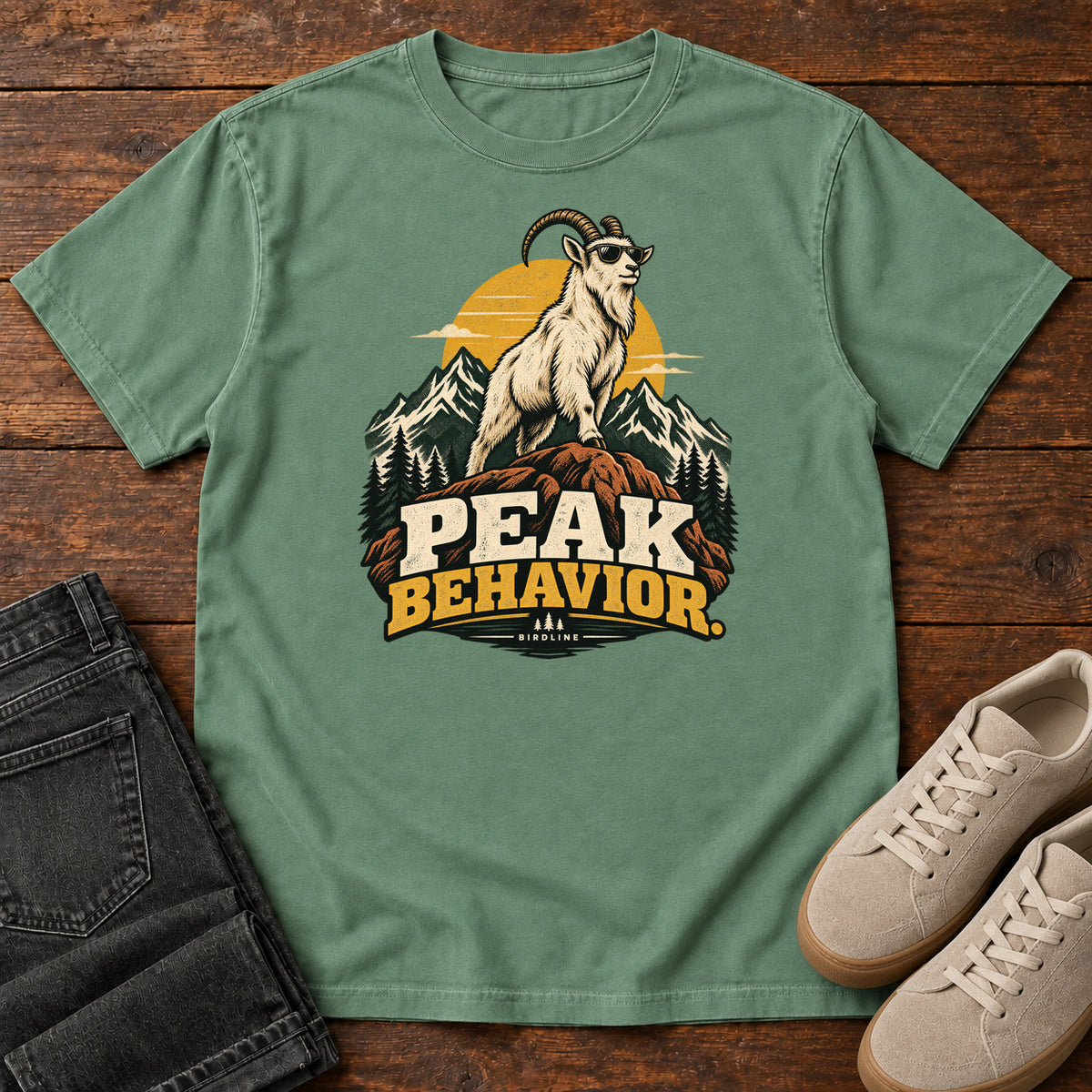 Peak Behavior T-Shirt in Light Green, front laid flat on wood, featuring a horned white goat wearing sunglasses on a rocky peak, with mountains, a gold sun and bold lettering.