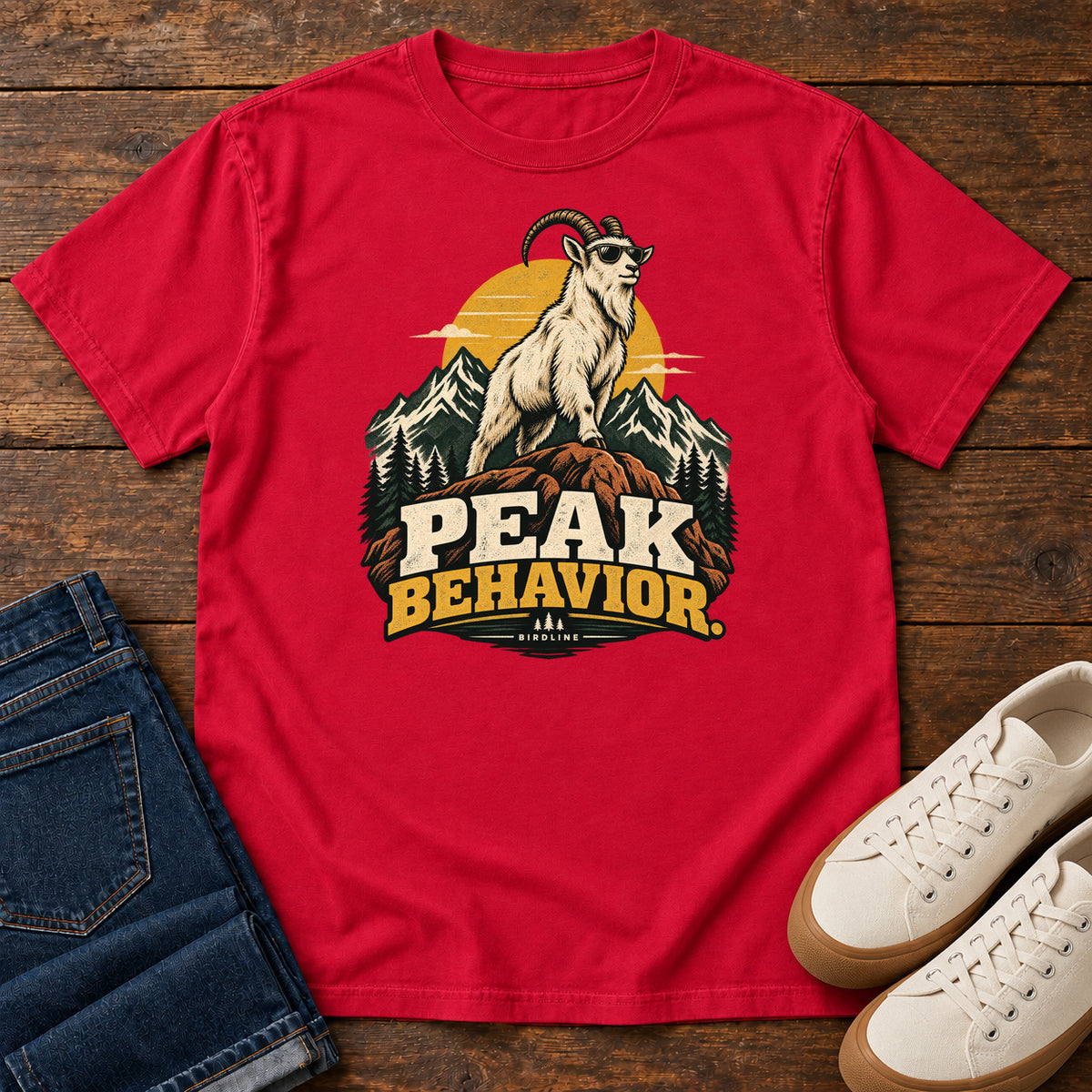 Peak Behavior T-Shirt in Red, front laid flat on wood, featuring a horned white goat wearing sunglasses on a rocky peak, with mountains, a gold sun and bold lettering.