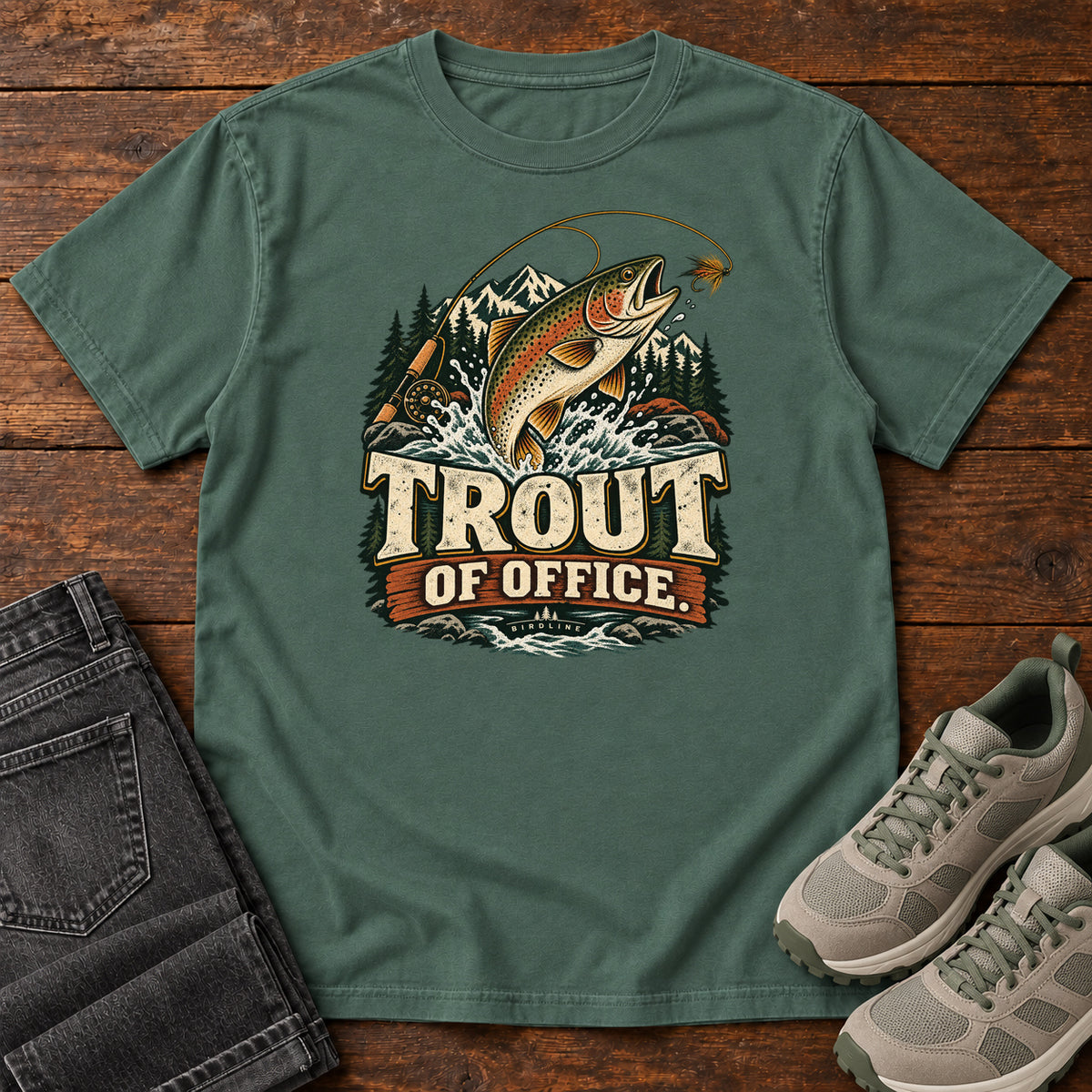 Blue Spruce Trout of Office Fly Rod T-Shirt with a spotted fish leaping toward a fishing fly, with mountains and cream lettering, front flat lay on wood.