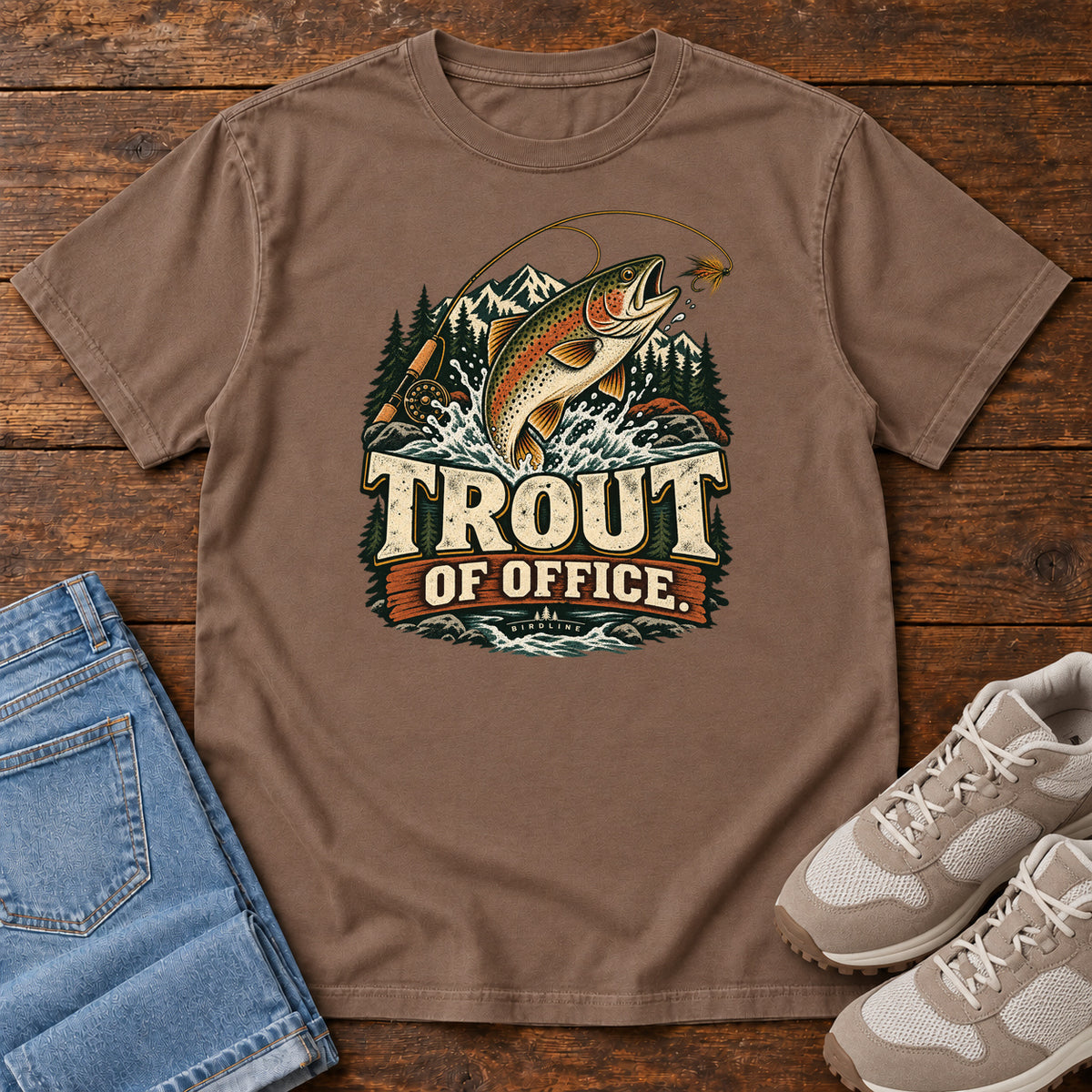 Espresso Trout of Office Fly Rod T-Shirt with a spotted fish leaping toward a fishing fly, with mountains and cream lettering, front flat lay on wood.