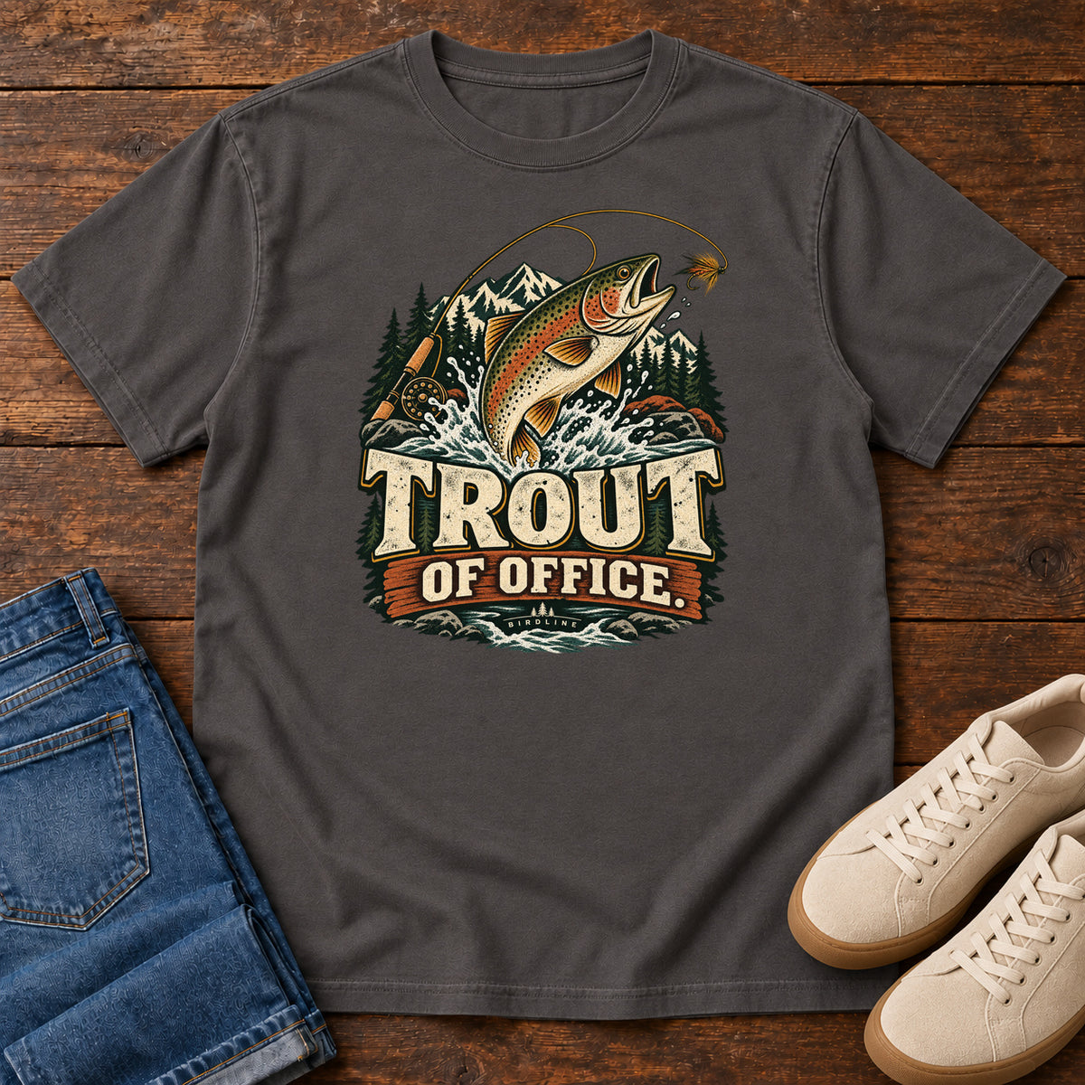 Graphite Trout of Office Fly Rod T-Shirt with a spotted fish leaping toward a fishing fly, with mountains and cream lettering, front flat lay on wood.