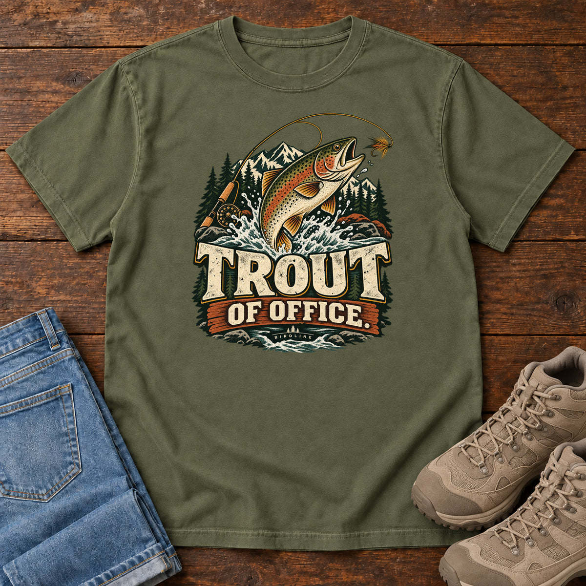 Hemp Trout of Office Fly Rod T-Shirt with a spotted fish leaping toward a fishing fly, with mountains and cream lettering, front flat lay on wood.