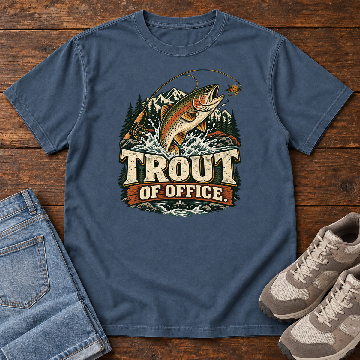 Midnight Trout of Office Fly Rod T-Shirt with a spotted fish leaping toward a fishing fly, with mountains and cream lettering, front flat lay on wood.