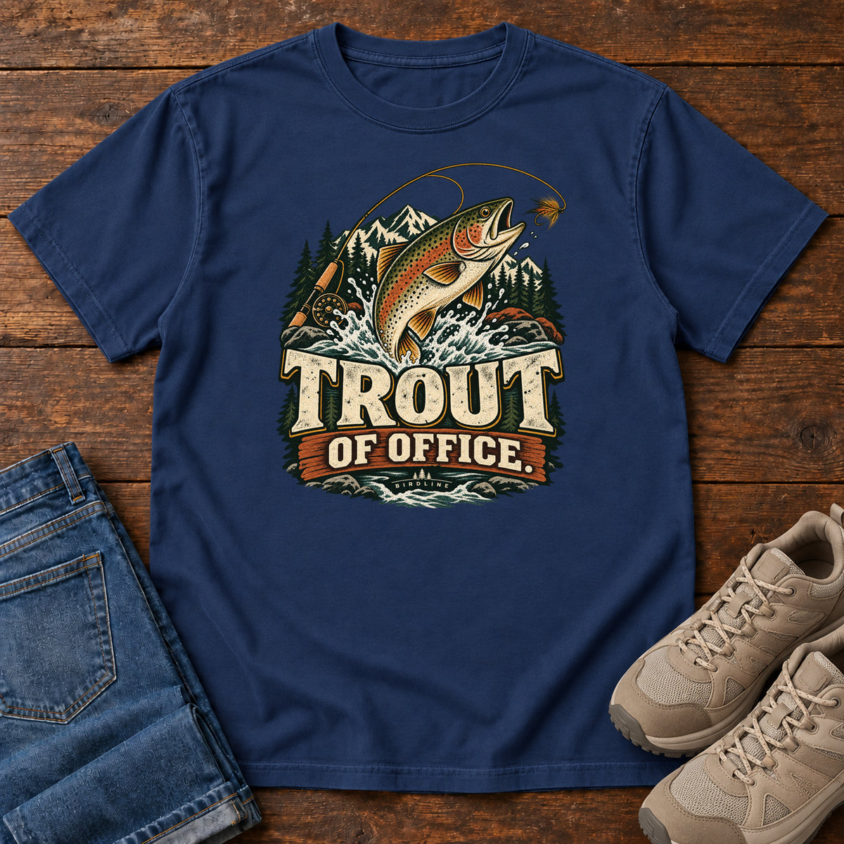 True Navy Trout of Office Fly Rod T-Shirt with a spotted fish leaping toward a fishing fly, with mountains and cream lettering, front flat lay on wood.