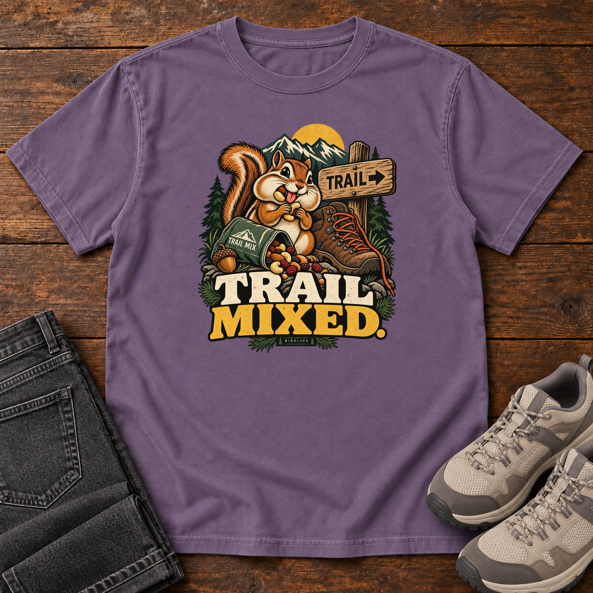 Grape Trail Mixed T-Shirt with a bushy-tailed cartoon animal beside a trail-mix bag, hiking boot and trail sign, front flat lay on wood.