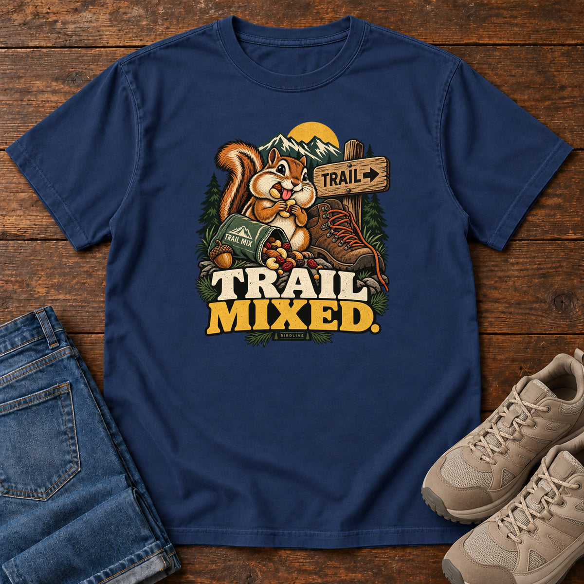 True Navy Trail Mixed T-Shirt with a bushy-tailed cartoon animal beside a trail-mix bag, hiking boot and trail sign, front flat lay on wood.