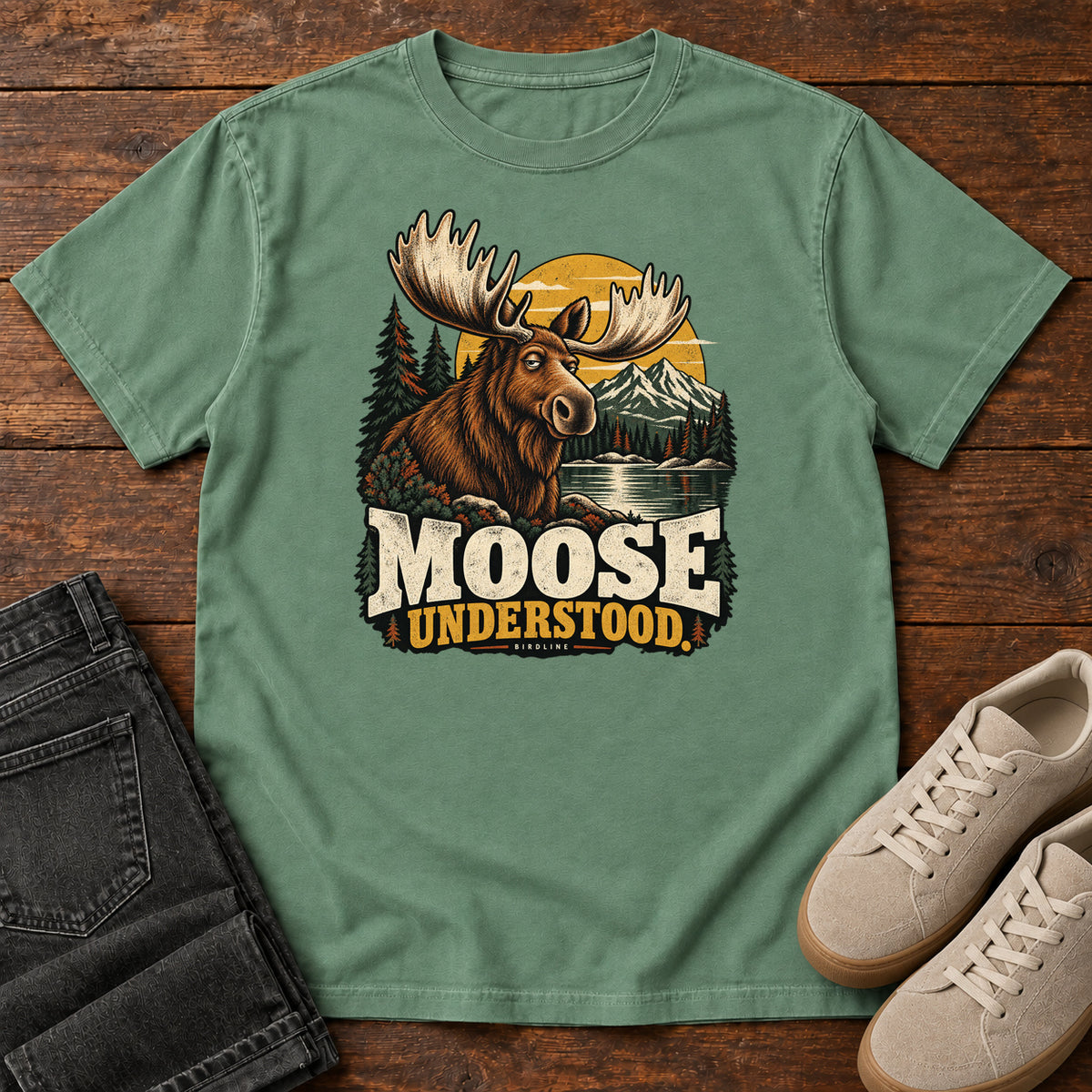 Light Green Moose Understood T-Shirt with an antlered moose beside a mountain lake, pines and a golden sun; front flat lay on wood.