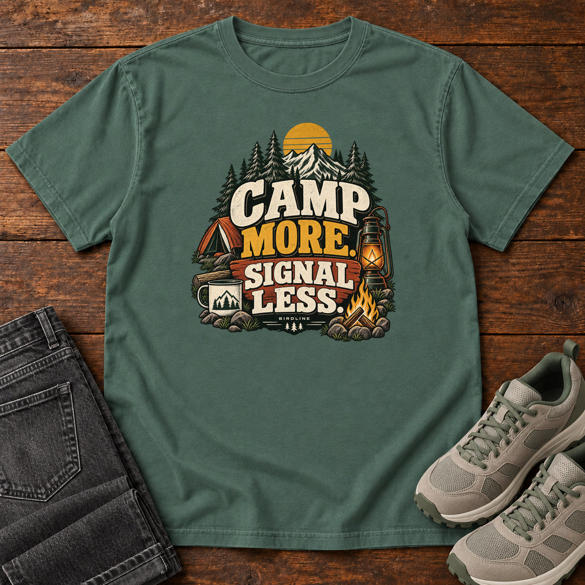 Blue Spruce Camp More Signal Less T-Shirt with a tent, lantern, campfire and mountain scene around bold slogan lettering; front flat lay on wood.