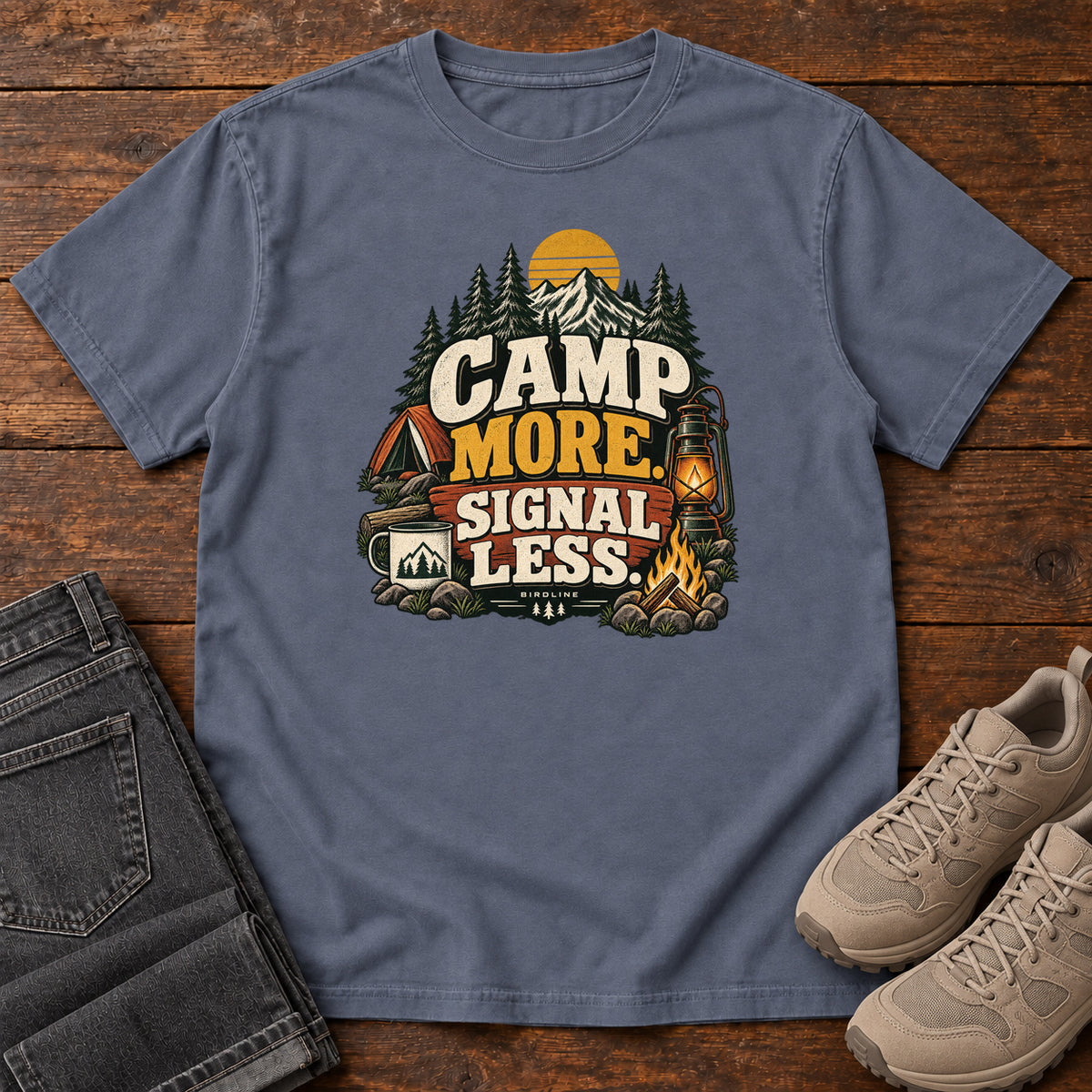 Denim Camp More Signal Less T-Shirt with a tent, lantern, campfire and mountain scene around bold slogan lettering; front flat lay on wood.