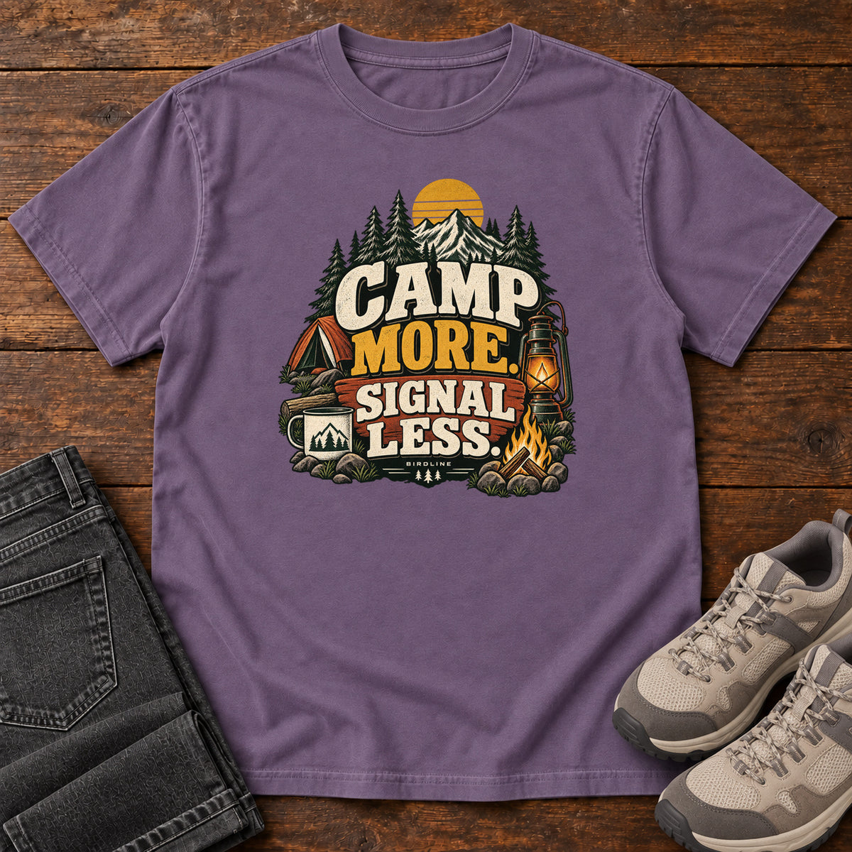 Grape Camp More Signal Less T-Shirt with a tent, lantern, campfire and mountain scene around bold slogan lettering; front flat lay on wood.