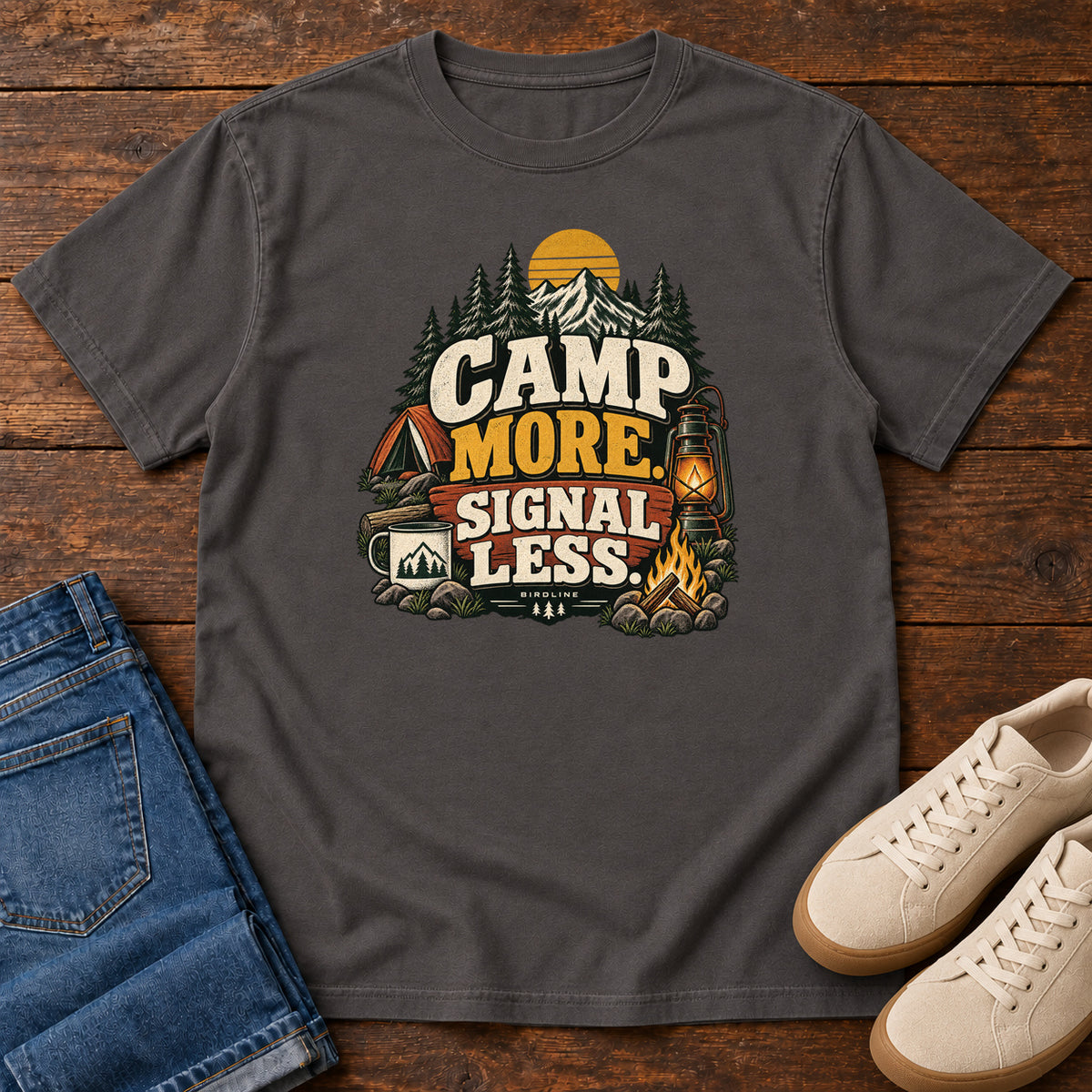Graphite Camp More Signal Less T-Shirt with a tent, lantern, campfire and mountain scene around bold slogan lettering; front flat lay on wood.