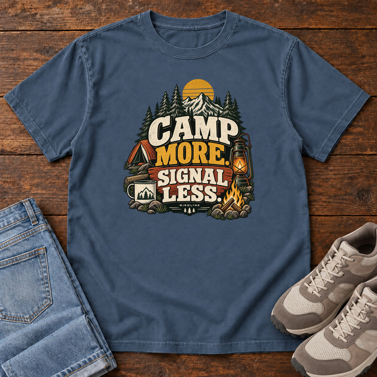 Midnight Camp More Signal Less T-Shirt with a tent, lantern, campfire and mountain scene around bold slogan lettering; front flat lay on wood.