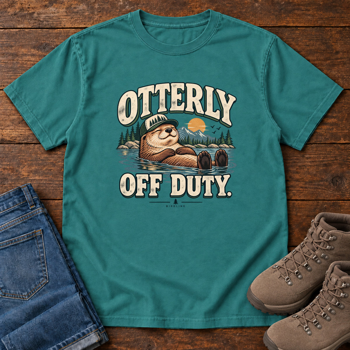 Emerald Otterly Off Duty T-Shirt with a floating otter in a knit cap, lake scenery and cream lettering, front flat lay on wood.