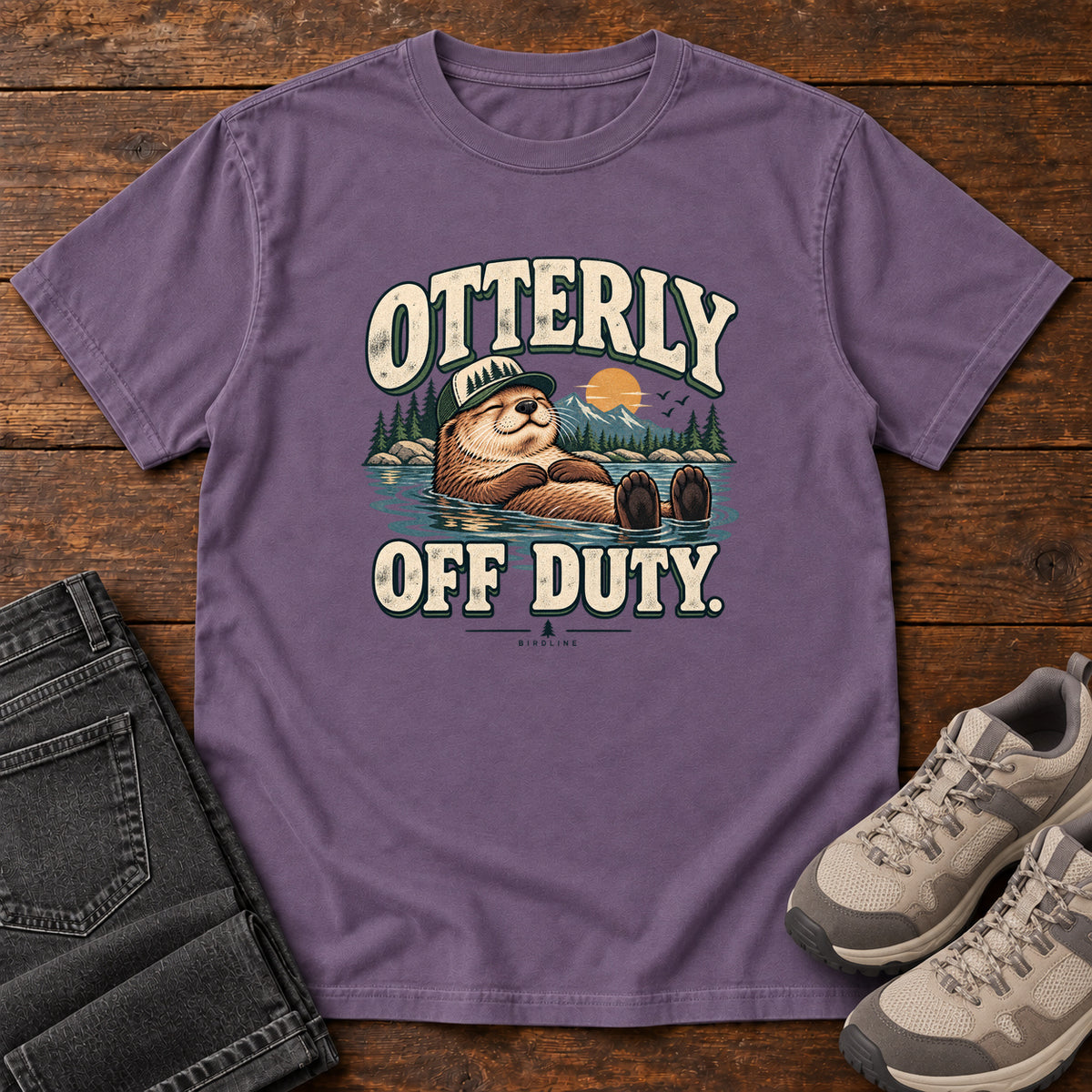 Grape Otterly Off Duty T-Shirt with a floating otter in a knit cap, lake scenery and cream lettering, front flat lay on wood.