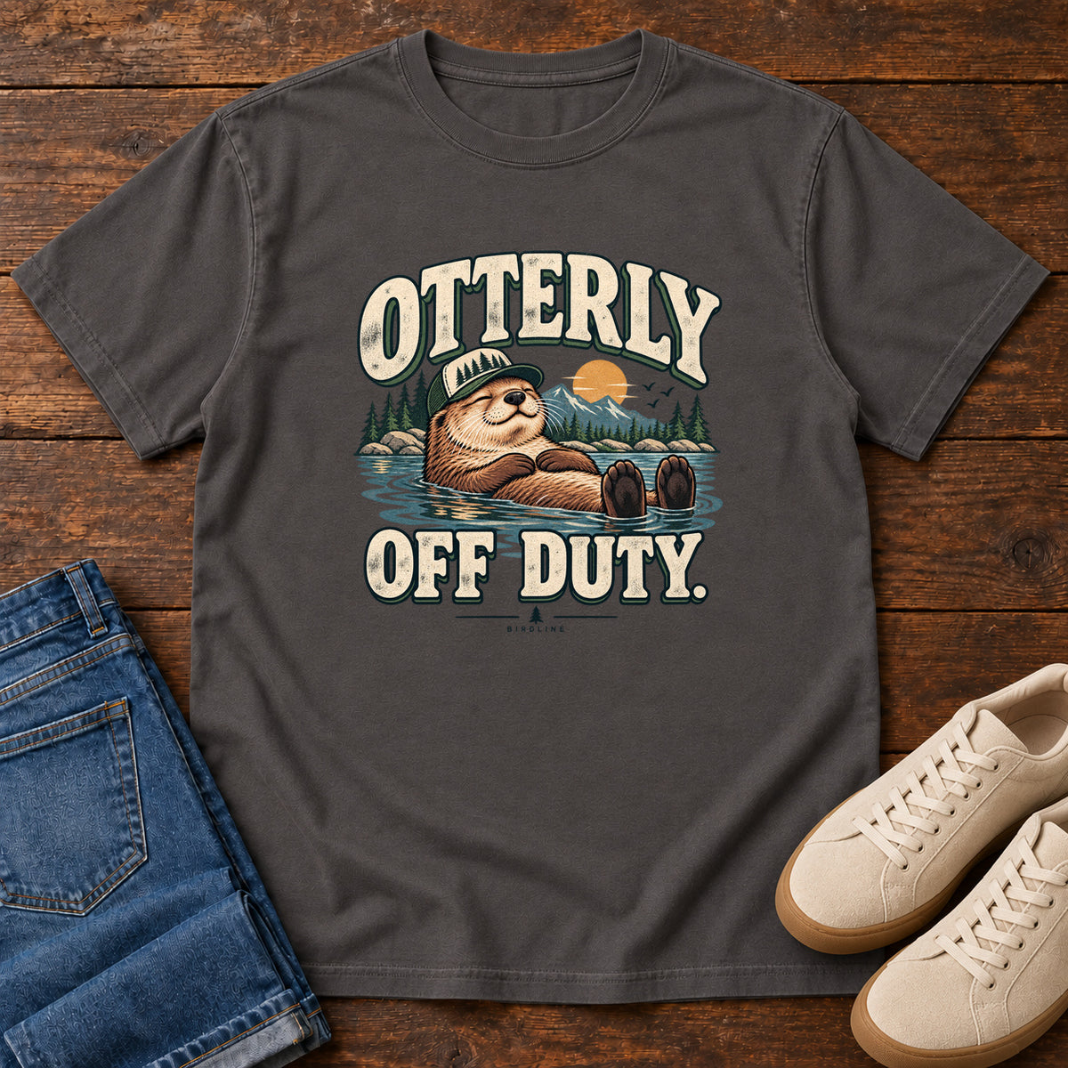 Graphite Otterly Off Duty T-Shirt with a floating otter in a knit cap, lake scenery and cream lettering, front flat lay on wood.