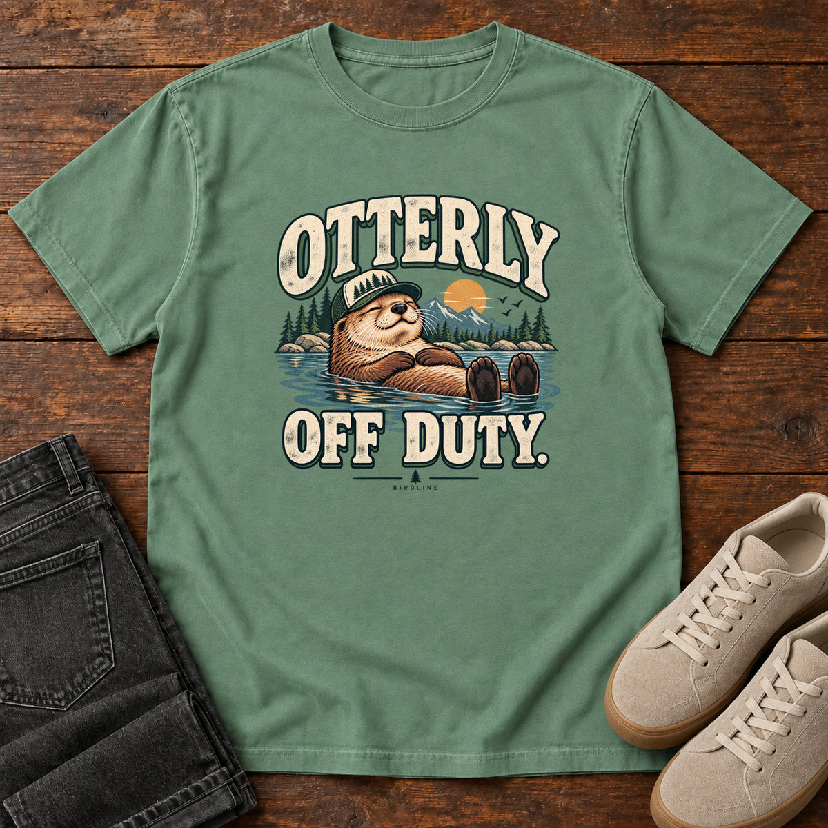 Light Green Otterly Off Duty T-Shirt with a floating otter in a knit cap, lake scenery and cream lettering, front flat lay on wood.