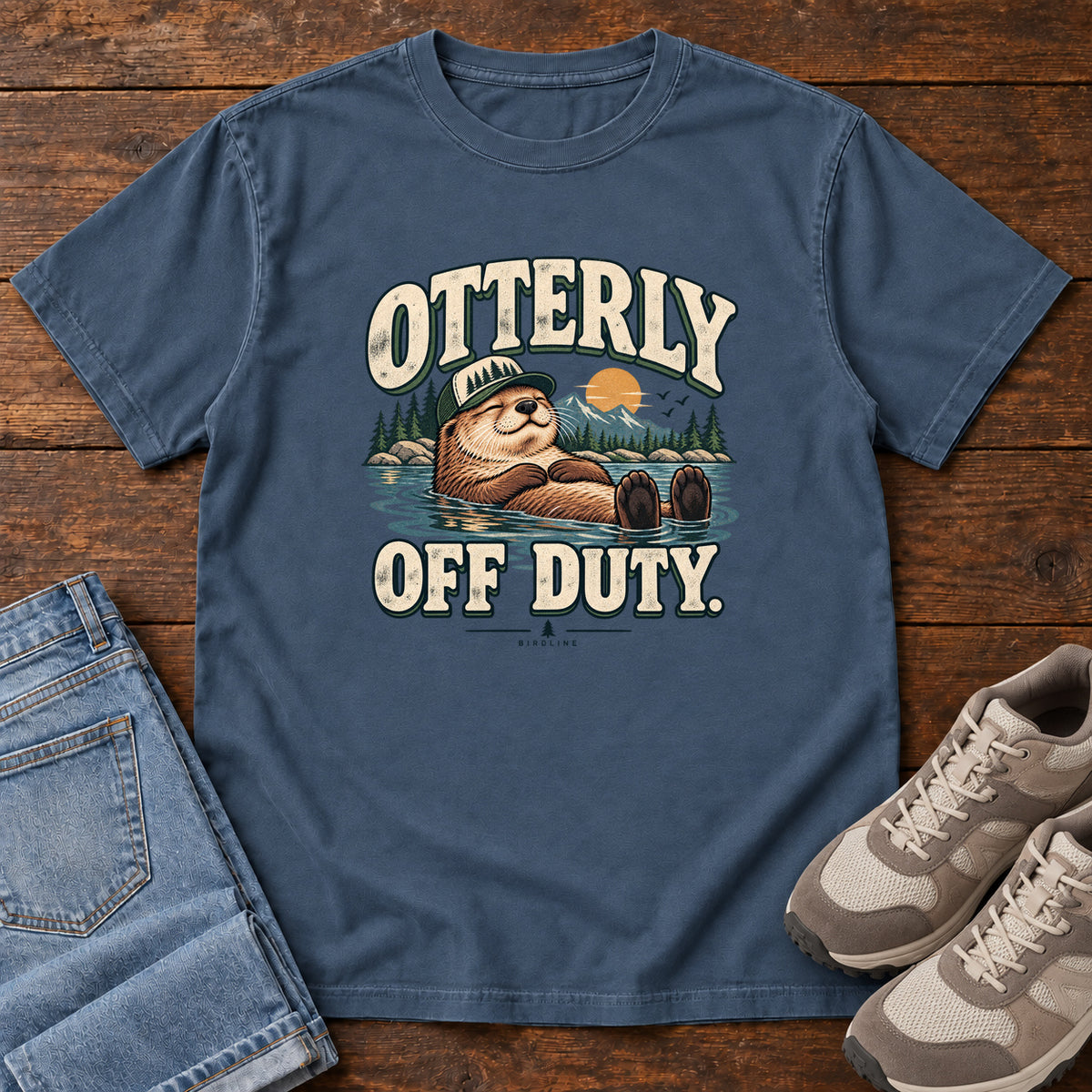 Midnight Otterly Off Duty T-Shirt with a floating otter in a knit cap, lake scenery and cream lettering, front flat lay on wood.