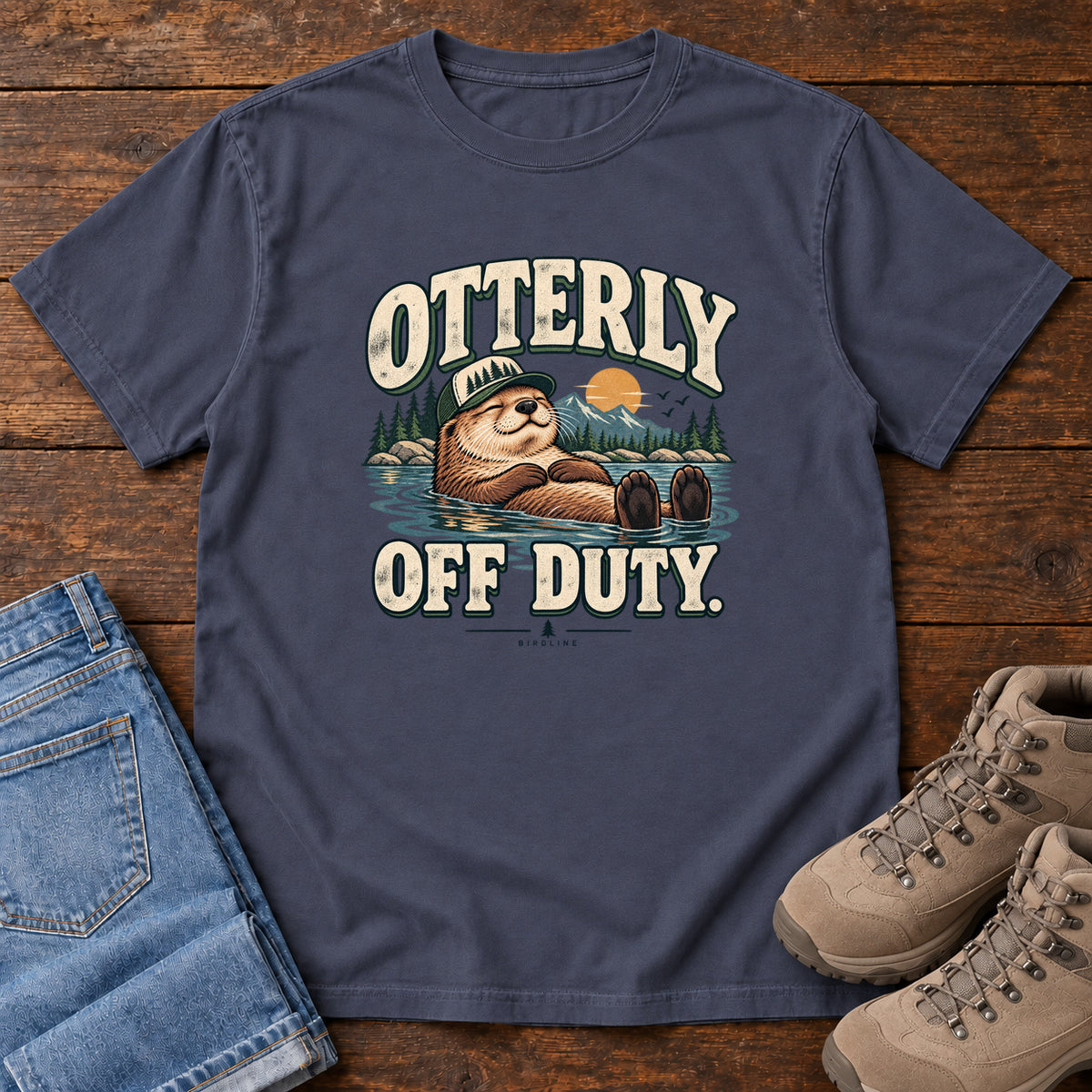 Navy Otterly Off Duty T-Shirt with a floating otter in a knit cap, lake scenery and cream lettering, front flat lay on wood.