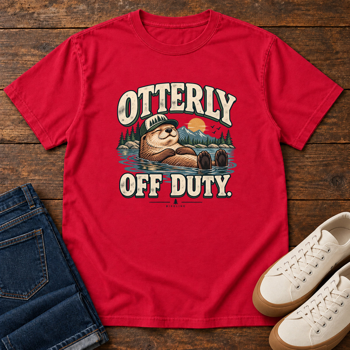 Red Otterly Off Duty T-Shirt with a floating otter in a knit cap, lake scenery and cream lettering, front flat lay on wood.