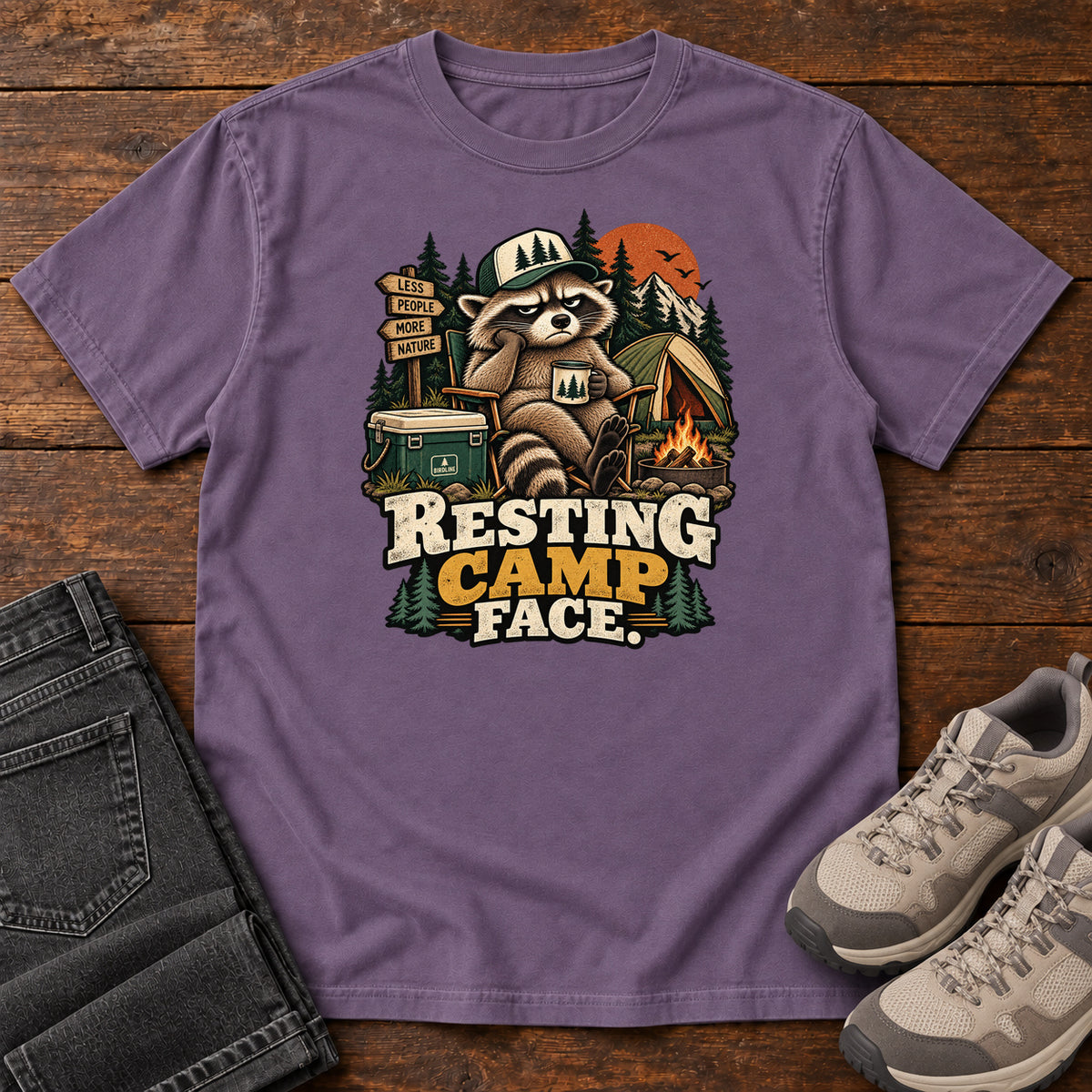 Resting Camp Face T-Shirt in Grape, front laid flat on wood, featuring a capped raccoon resting in a camp chair with a mug beside a tent, cooler and campfire.