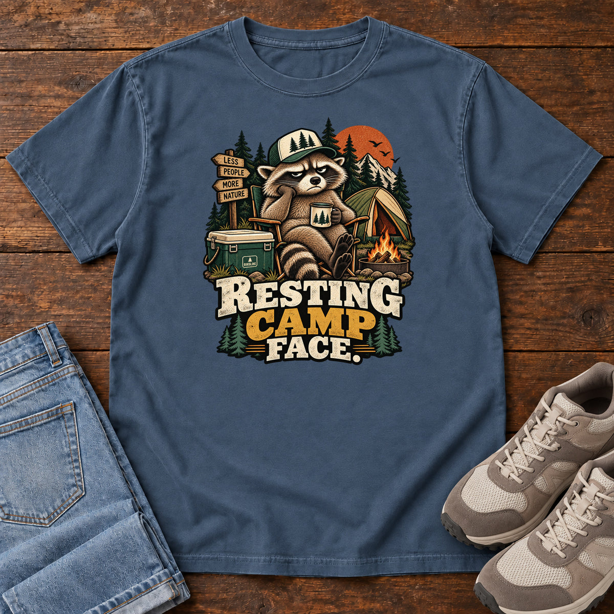 Resting Camp Face T-Shirt in Midnight, front laid flat on wood, featuring a capped raccoon resting in a camp chair with a mug beside a tent, cooler and campfire.