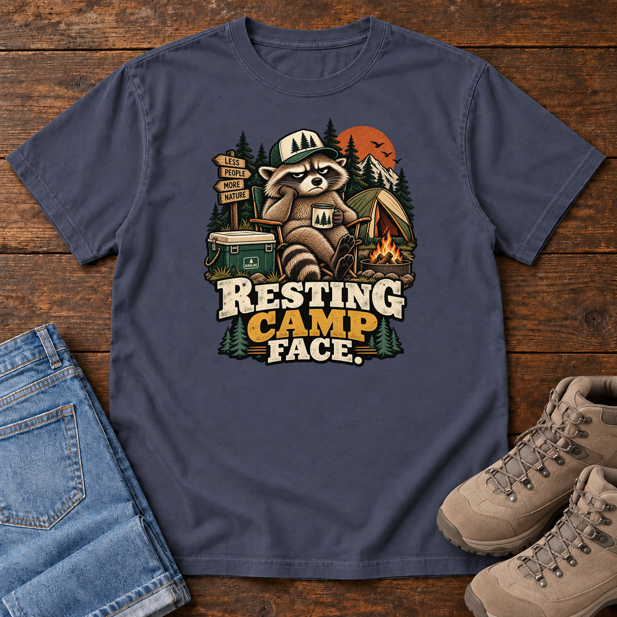 Resting Camp Face T-Shirt in Navy, front laid flat on wood, featuring a capped raccoon resting in a camp chair with a mug beside a tent, cooler and campfire.