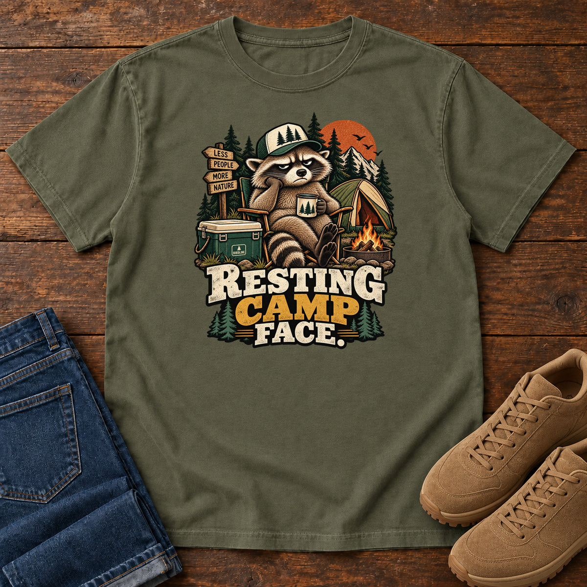 Resting Camp Face T-Shirt in Sage, front laid flat on wood, featuring a capped raccoon resting in a camp chair with a mug beside a tent, cooler and campfire.