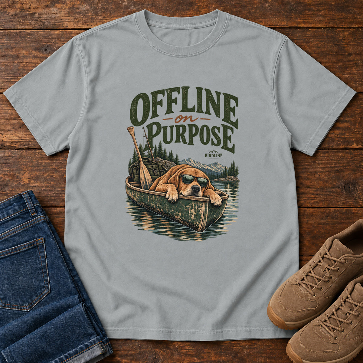 Granite Offline on Purpose Canoe T-Shirt with a brown dog wearing sunglasses and resting in a canoe below dark green lettering, front flat lay on wood.