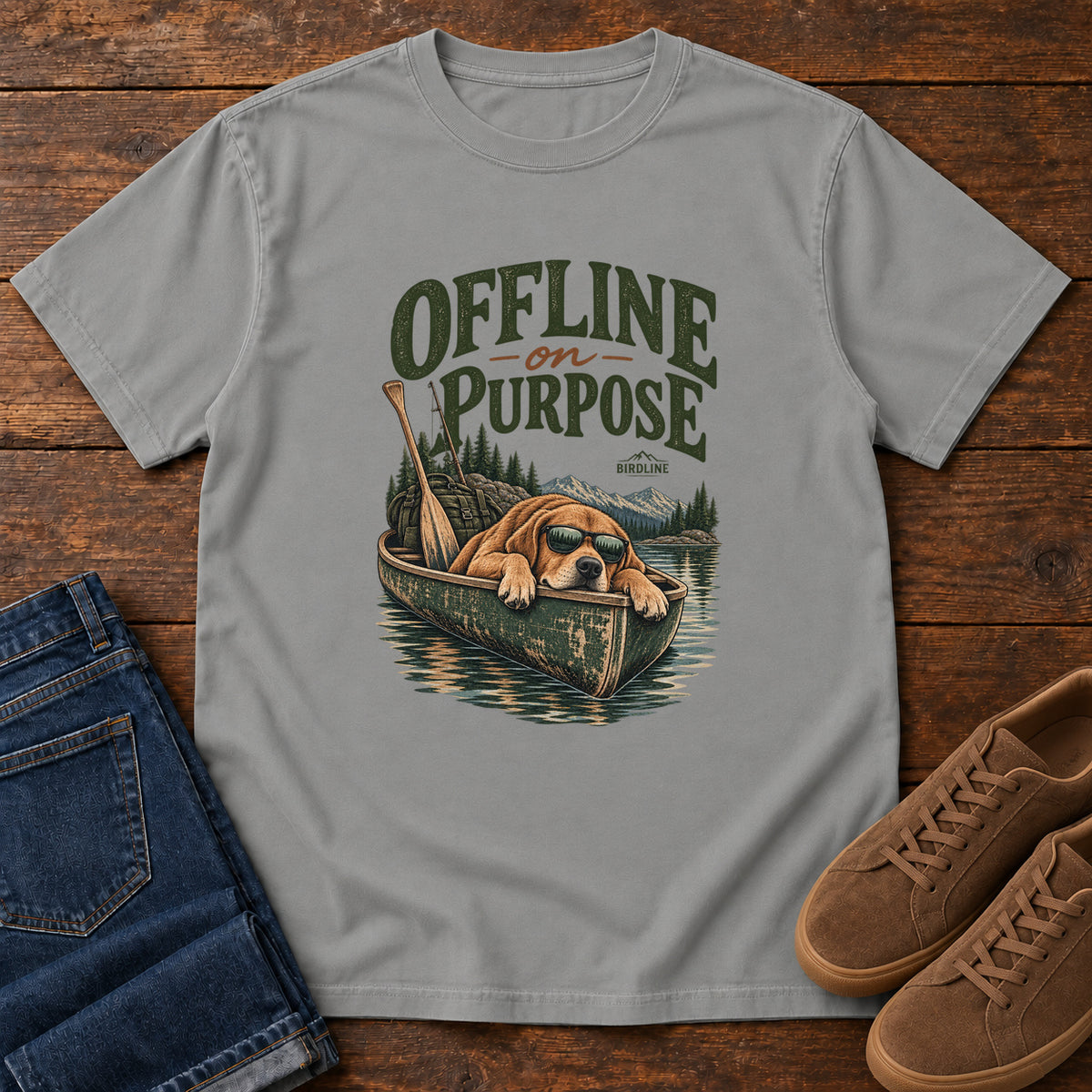 Grey Offline on Purpose Canoe T-Shirt with a brown dog wearing sunglasses and resting in a canoe below dark green lettering, front flat lay on wood.