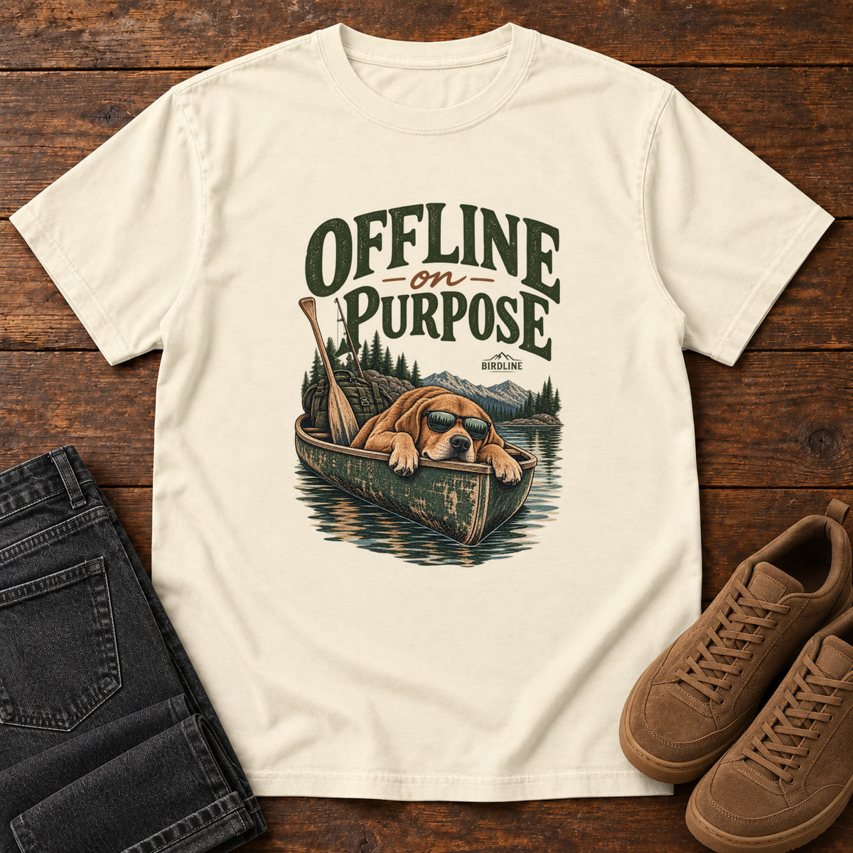 Ivory Offline on Purpose Canoe T-Shirt with a brown dog wearing sunglasses and resting in a canoe below dark green lettering, front flat lay on wood.