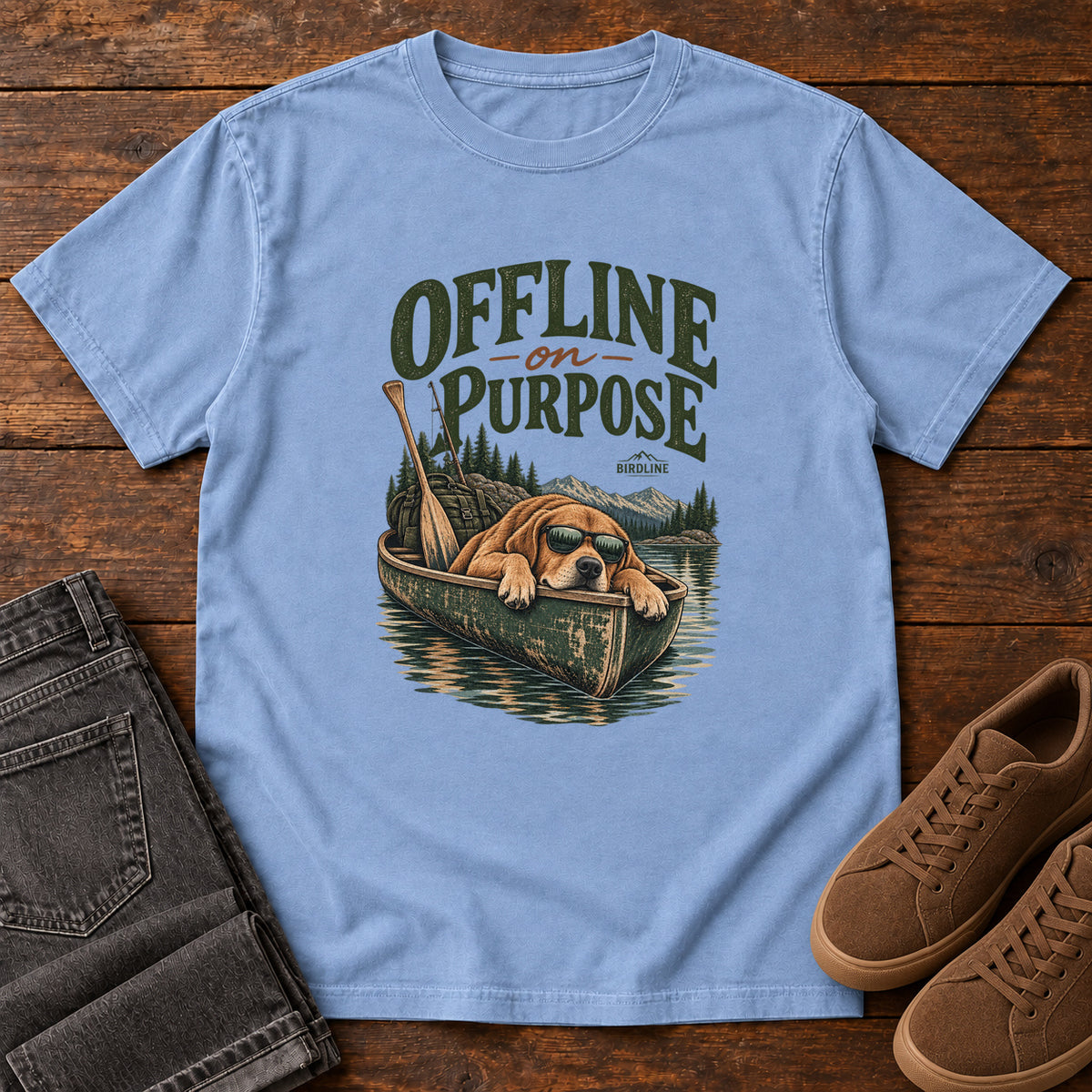 Washed Denim Offline on Purpose Canoe T-Shirt with a brown dog wearing sunglasses and resting in a canoe below dark green lettering, front flat lay on wood.