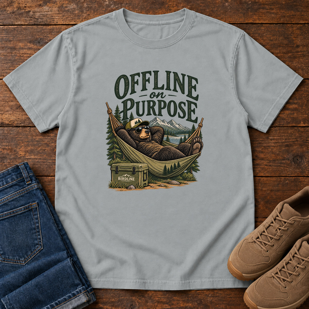 Granite Offline on Purpose T-Shirt with a bear in sunglasses relaxing in a hammock beside a cooler and mountain lake; front flat lay on wood.