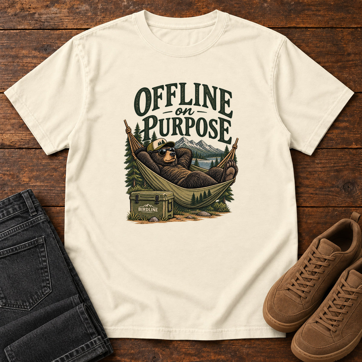 Ivory Offline on Purpose T-Shirt with a bear in sunglasses relaxing in a hammock beside a cooler and mountain lake; front flat lay on wood.