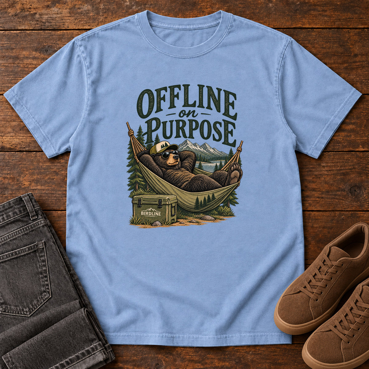 Washed Denim Offline on Purpose T-Shirt with a bear in sunglasses relaxing in a hammock beside a cooler and mountain lake; front flat lay on wood.