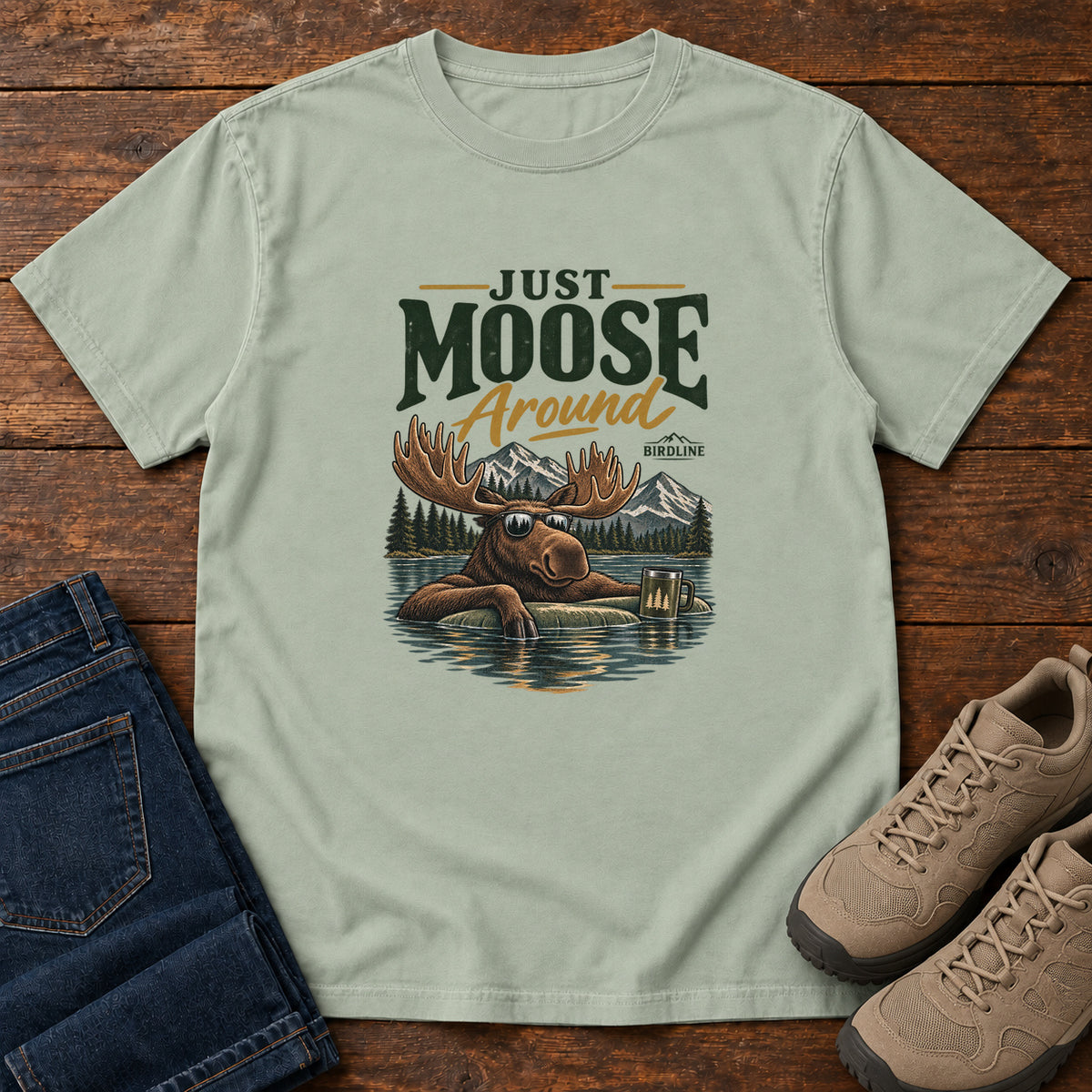 Bay Just Moose Around T-Shirt with a moose wearing sunglasses in a lake beneath mountains and slogan lettering; front flat lay on wood.