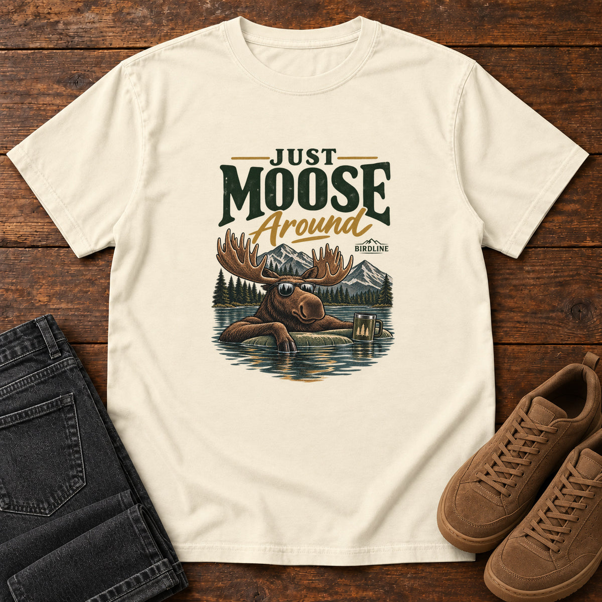 Ivory Just Moose Around T-Shirt with a moose wearing sunglasses in a lake beneath mountains and slogan lettering; front flat lay on wood.