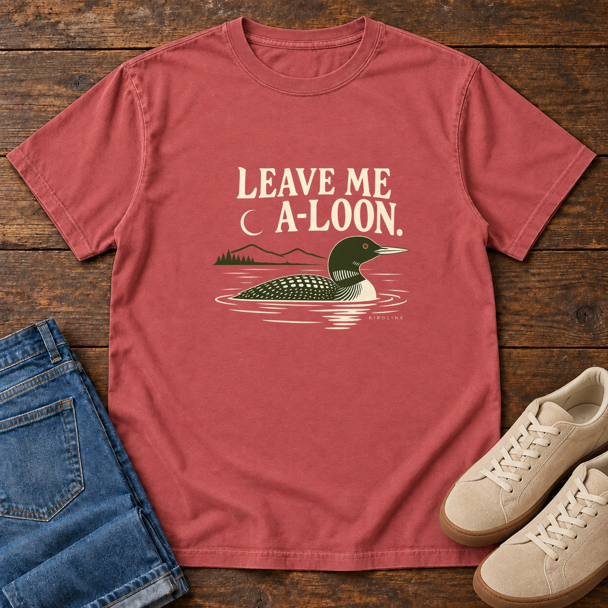 Crimson Leave Me A Loon T-Shirt with a green-and-cream waterbird floating below a crescent moon and cream lettering, front flat lay on wood.
