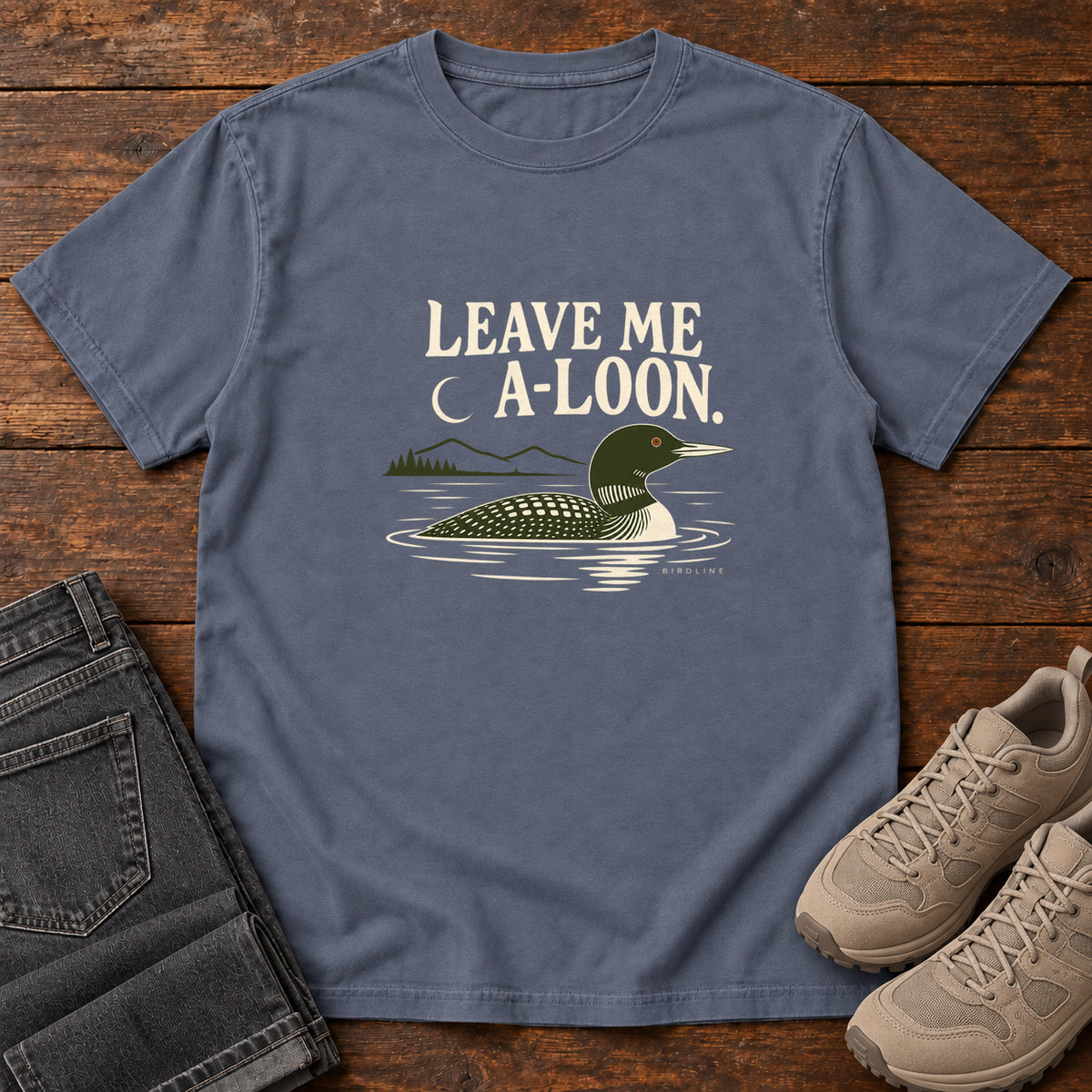 Denim Leave Me A Loon T-Shirt with a green-and-cream waterbird floating below a crescent moon and cream lettering, front flat lay on wood.