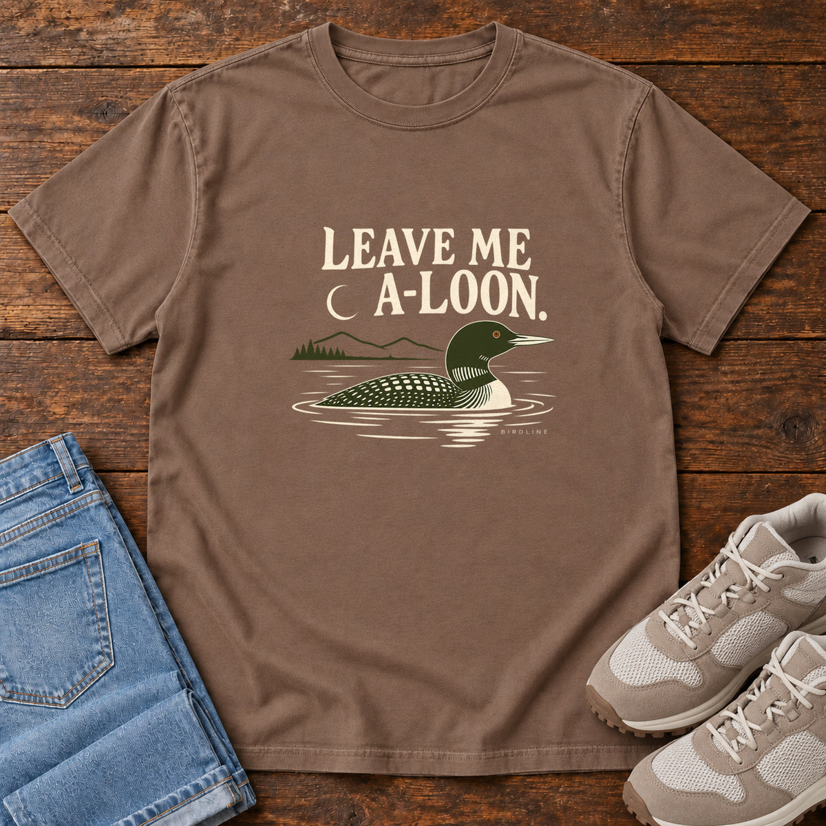 Espresso Leave Me A Loon T-Shirt with a green-and-cream waterbird floating below a crescent moon and cream lettering, front flat lay on wood.