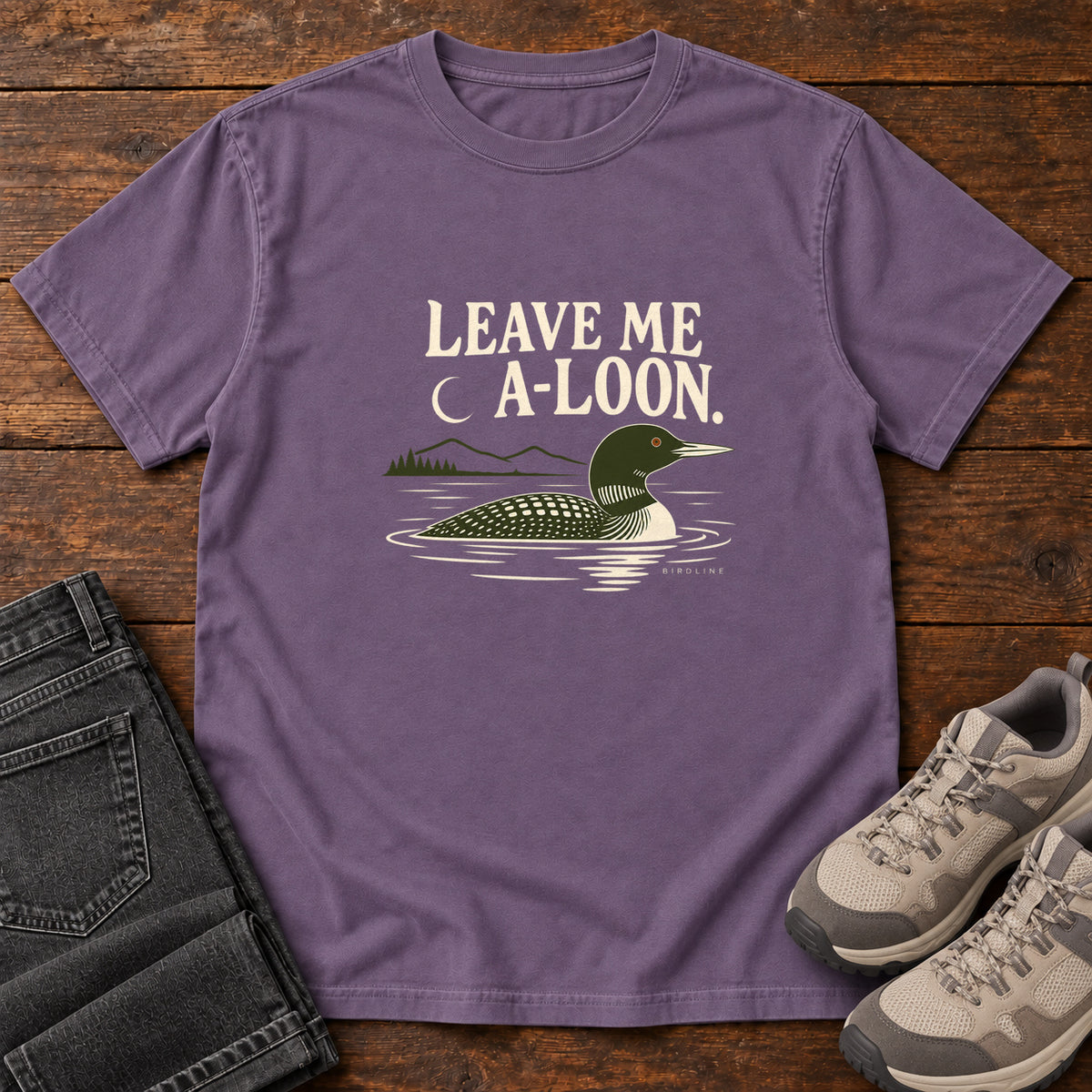 Grape Leave Me A Loon T-Shirt with a green-and-cream waterbird floating below a crescent moon and cream lettering, front flat lay on wood.