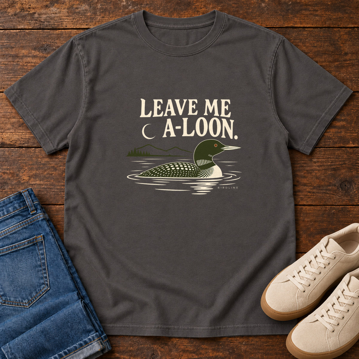 Graphite Leave Me A Loon T-Shirt with a green-and-cream waterbird floating below a crescent moon and cream lettering, front flat lay on wood.