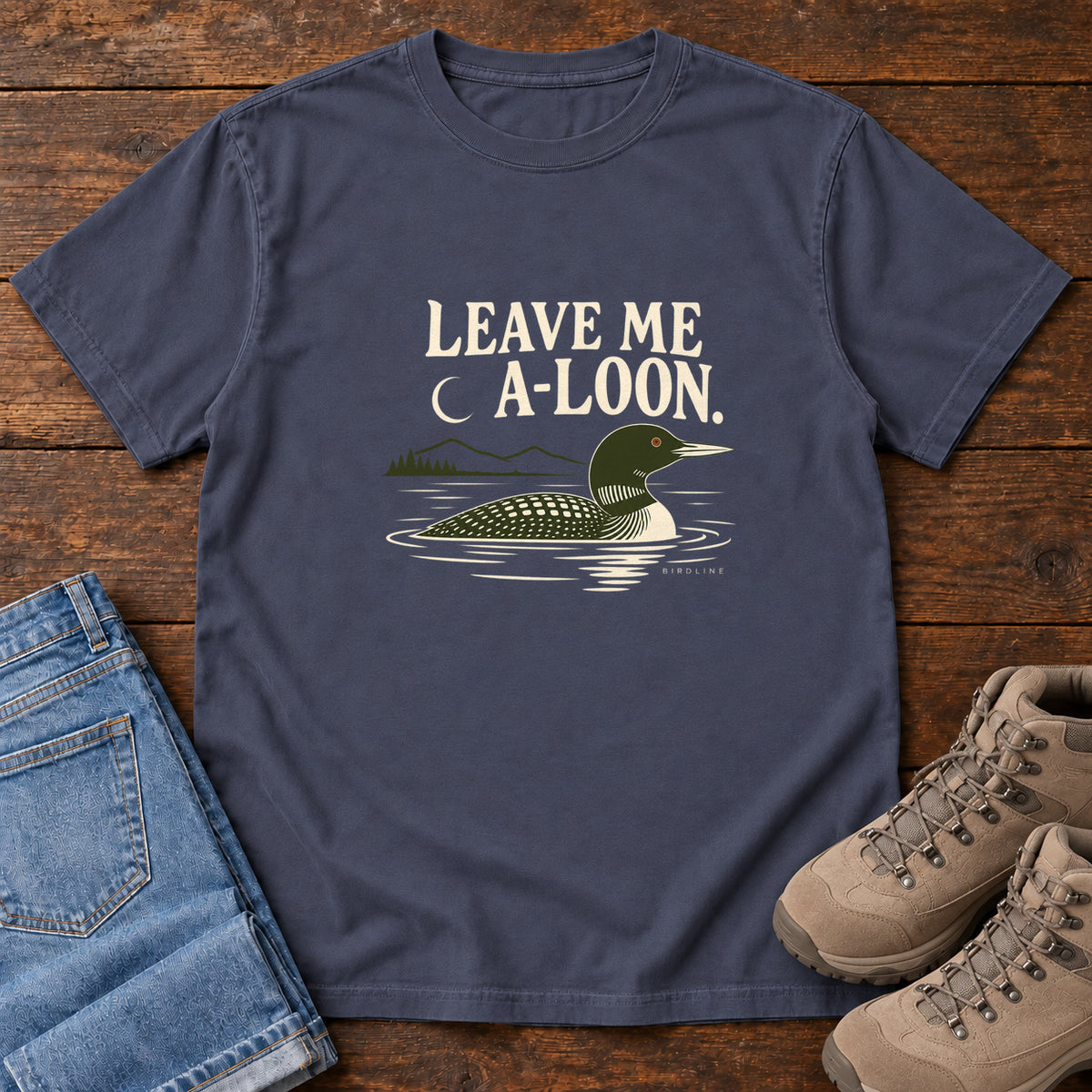 Navy Leave Me A Loon T-Shirt with a green-and-cream waterbird floating below a crescent moon and cream lettering, front flat lay on wood.